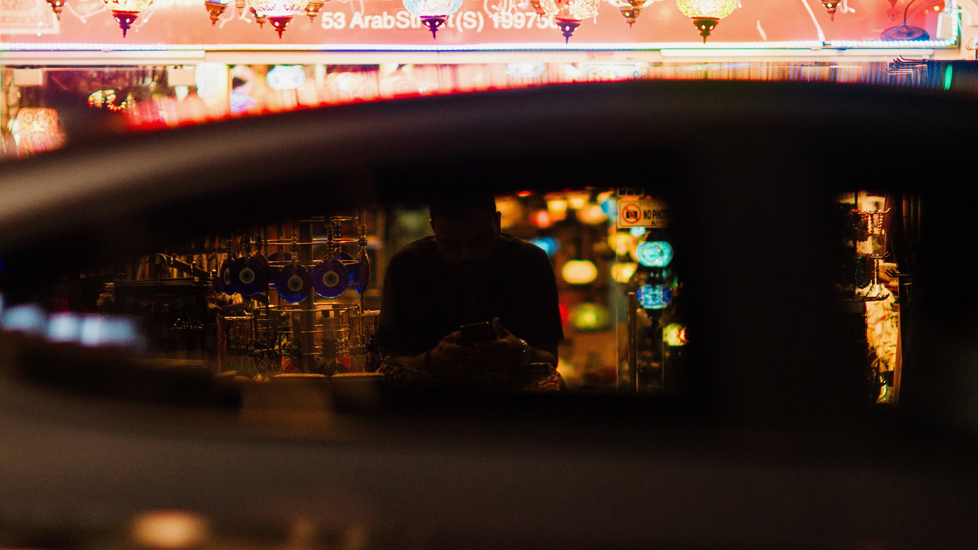 A high-fidelity 16:9 cinematic unit still of Arab Street, Singapore at night. Captured through a reflective foreground layer to create a voyeuristic, nocturnal narrative. A silhouetted figure is framed by glowing, intricate lanterns, utilizing a high-contrast noir aesthetic with deep shadows and warm ambient highlights.