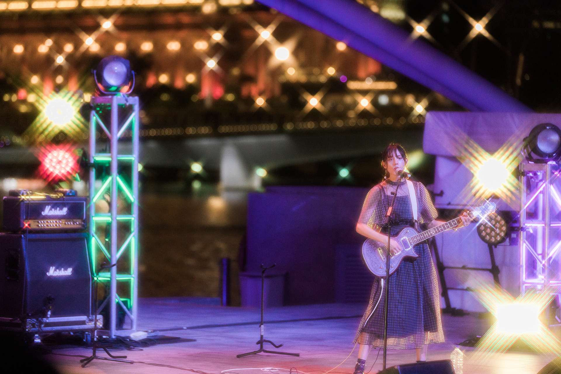Cinematic unit-still of a live musical performance at night; high-fidelity capture of stage lighting, starburst lens flares, and technical rigging with an urban waterfront backdrop