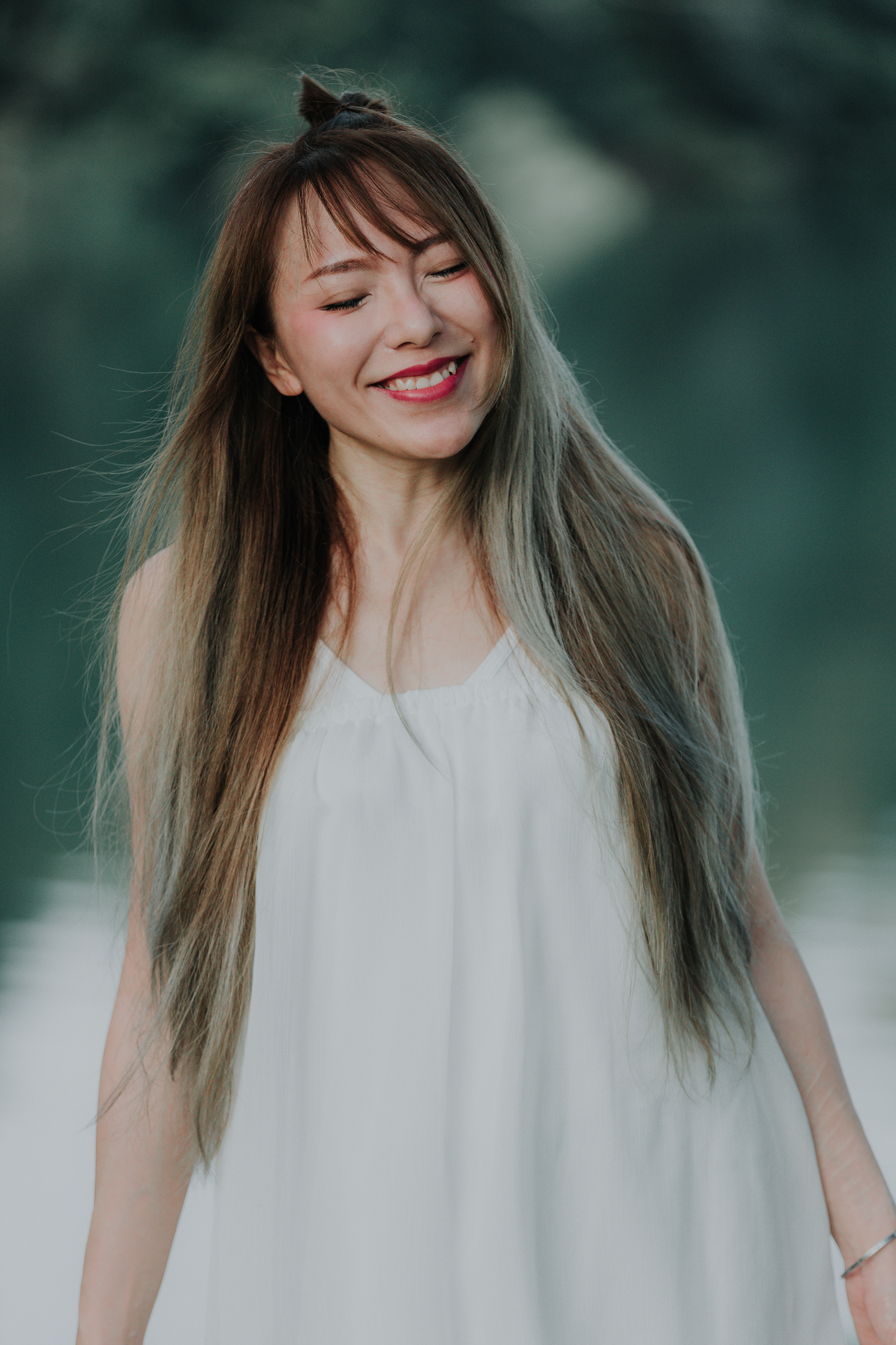 Close-up candid modern portrait of a woman in a white dress with long hair, smiling with her eyes closed in natural outdoor light.