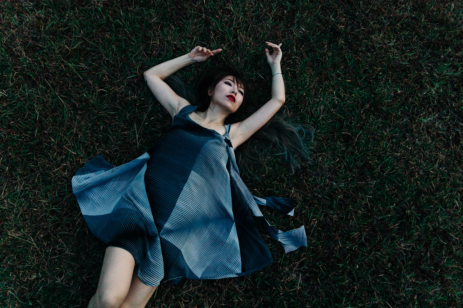 Vertical top-down (overheard) photograph of a woman with red lipstick and a geometric blue dress lying dramatically in dark green grass.
