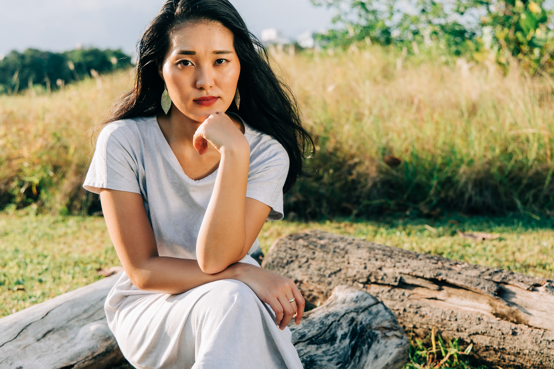 Horizontal outdoor portrait of a woman in a light-colored dress sitting on a log, resting her chin on her hand, with sunset light, water, and distant trees.