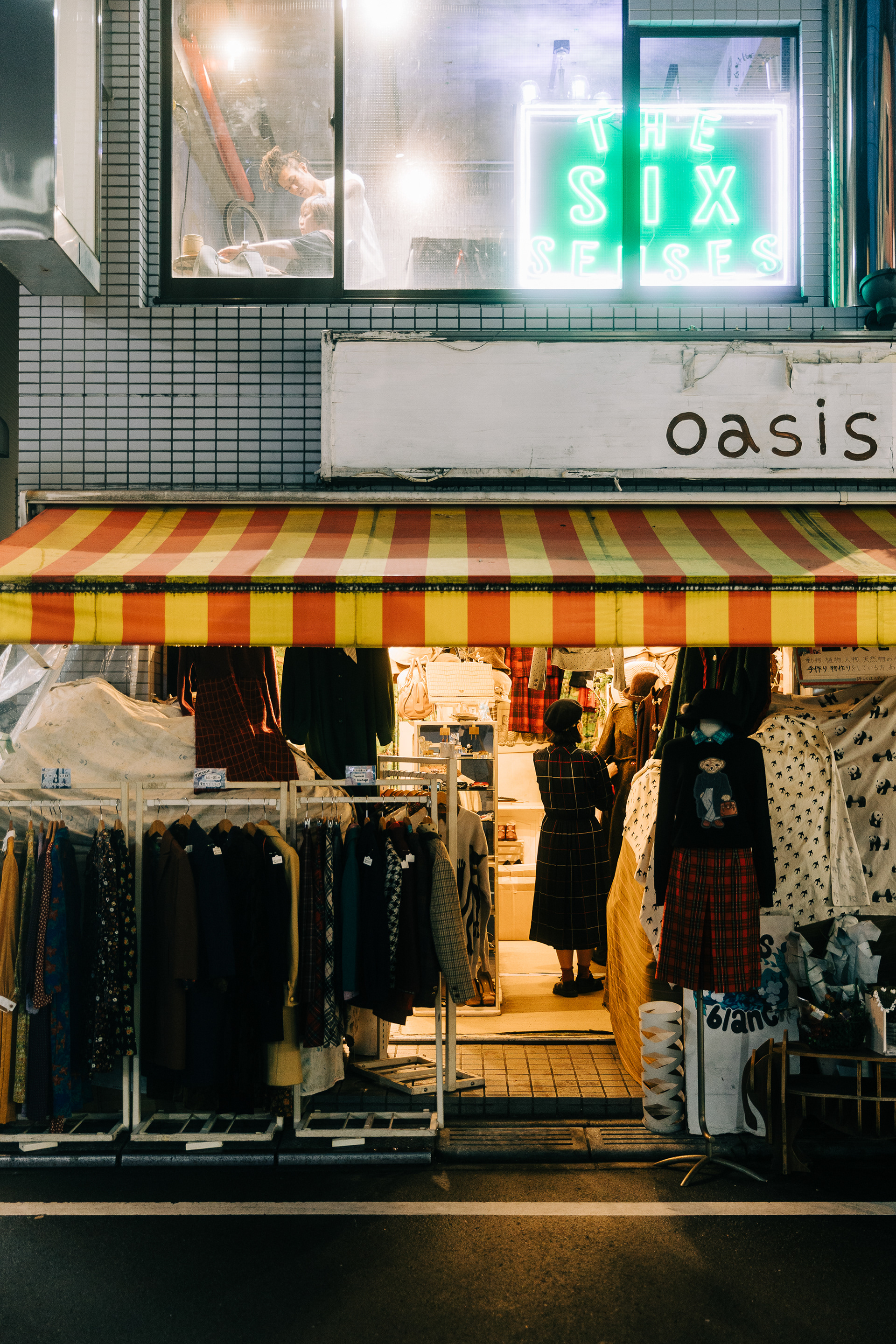 Atmospheric night shot of a Tokyo storefront named "Oasis" with a striped awning. Overlapping green neon signage and warm interior lighting create high visual friction.