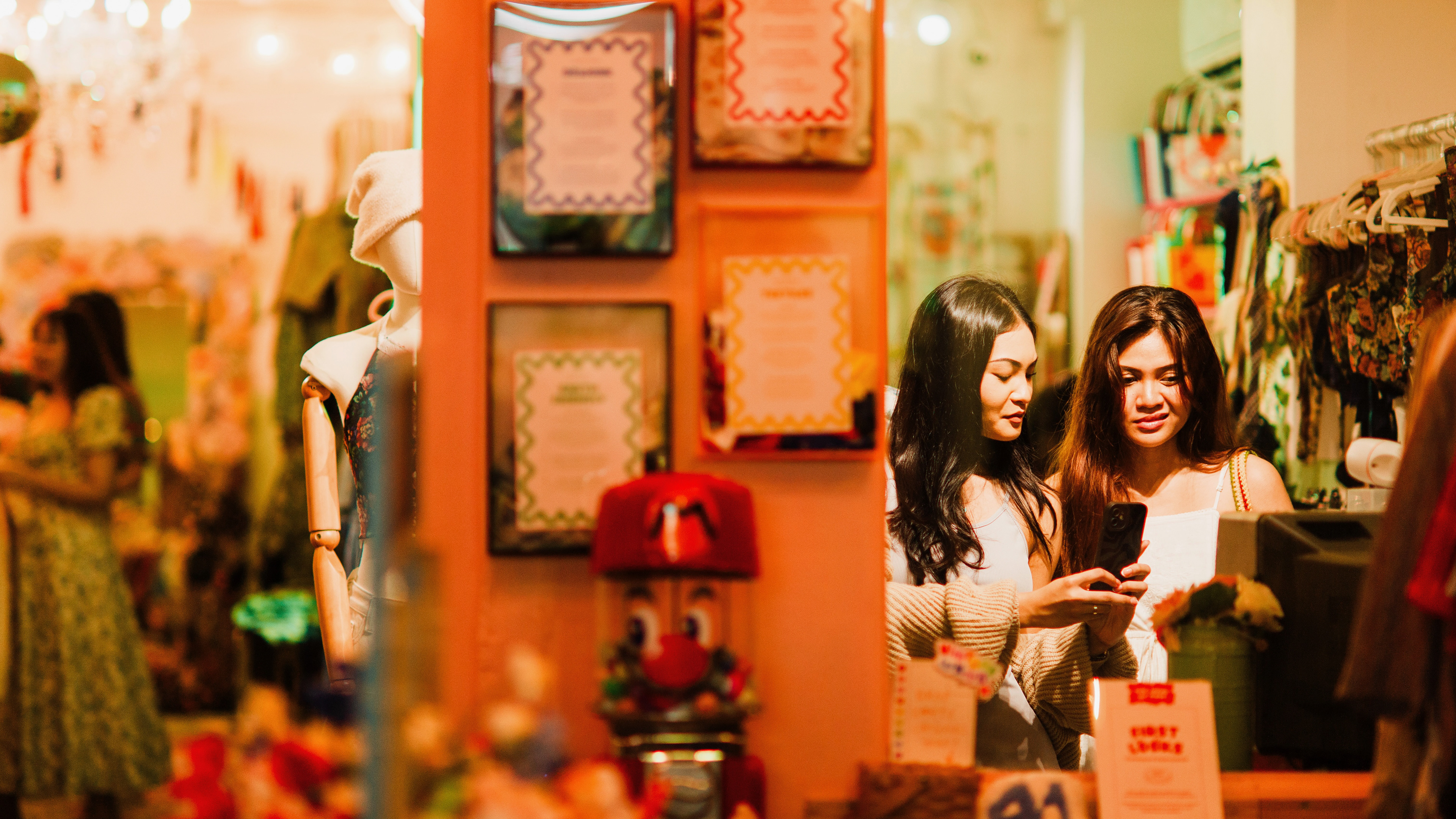 A 16:9 cinematic unit still capturing two women in a warmly lit, boutique environment in Singapore. Strategic use of a central orange pillar to create a voyeuristic split-frame composition. High-fidelity color grading with warm peach tones and industrial green accents. A modern, candid narrative study in nocturnal urban life.