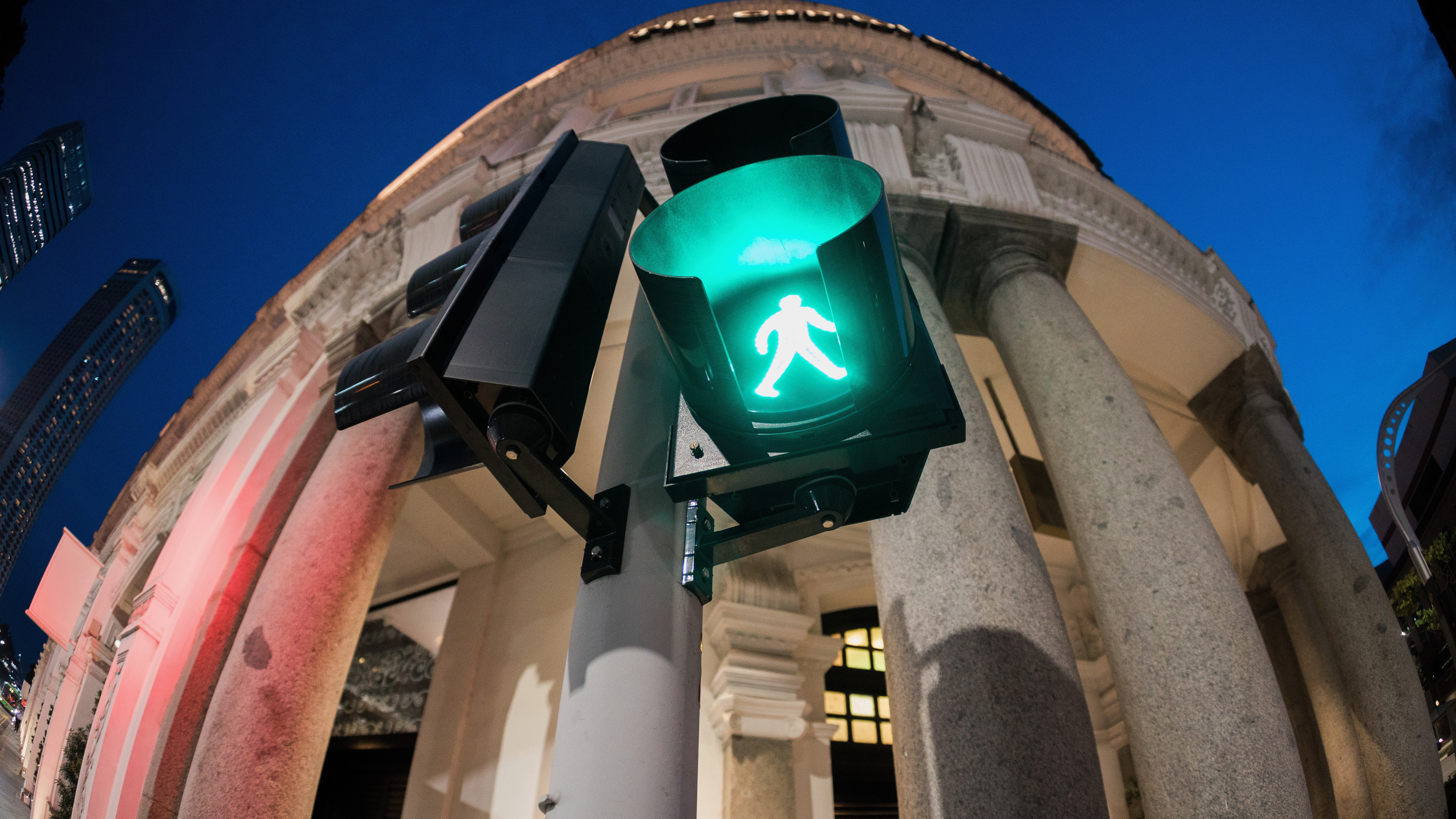 A cinematic low-angle study of a green pedestrian signal in Singapore. Fisheye perspective capturing the neoclassical architecture of the background against a deep blue dusk sky. A mechanical noir aesthetic focusing on urban light refractions and architectural distortion.