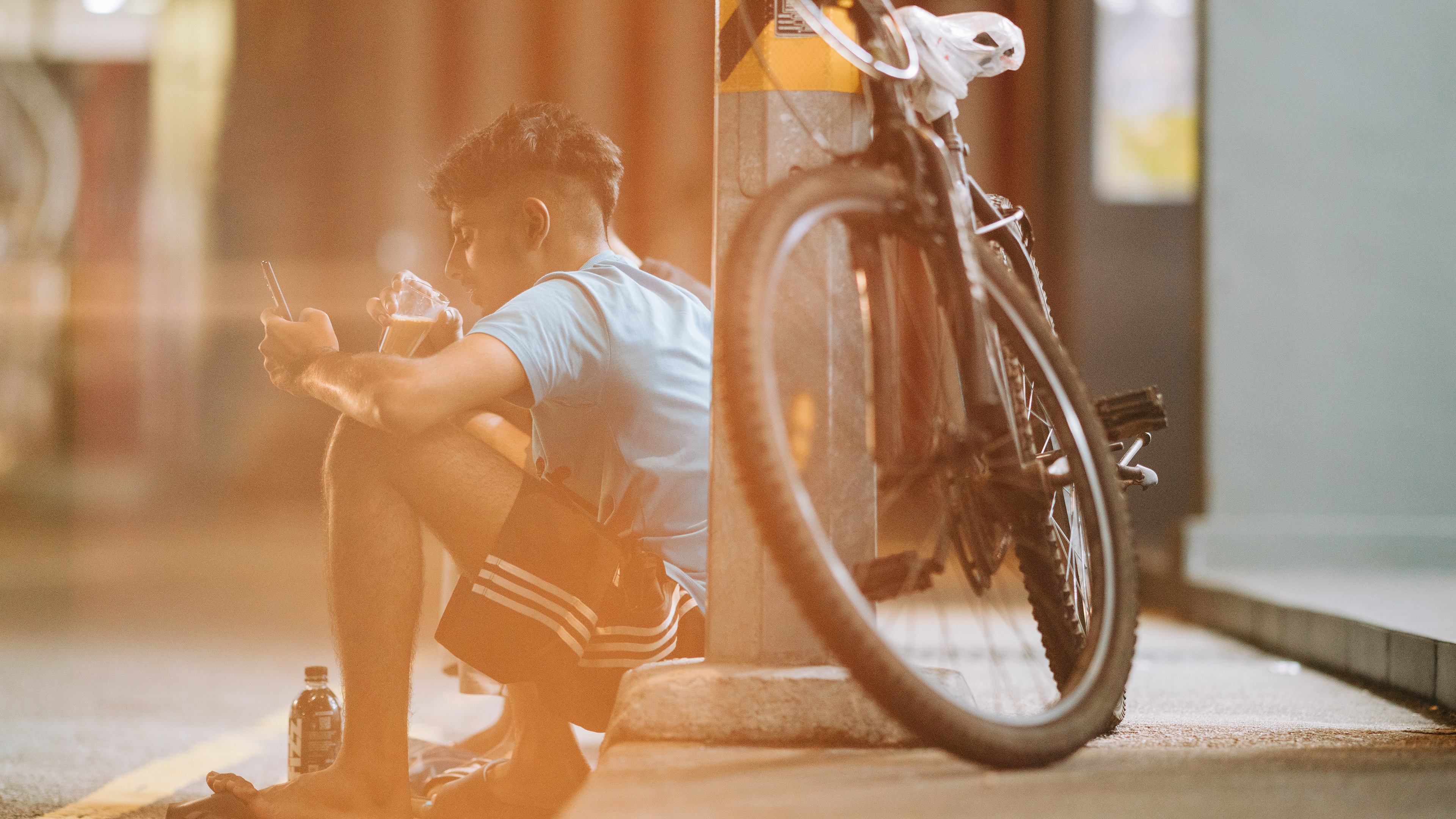 A 16:9 cinematic unit still of a young man resting on a curb in Singapore. Captured with a warm, golden-hour flare and a voyeuristic perspective through a bicycle wheel. A high-fidelity study in urban life, candid humanity, and the soft side of the "Mechanical Soul" aesthetic.