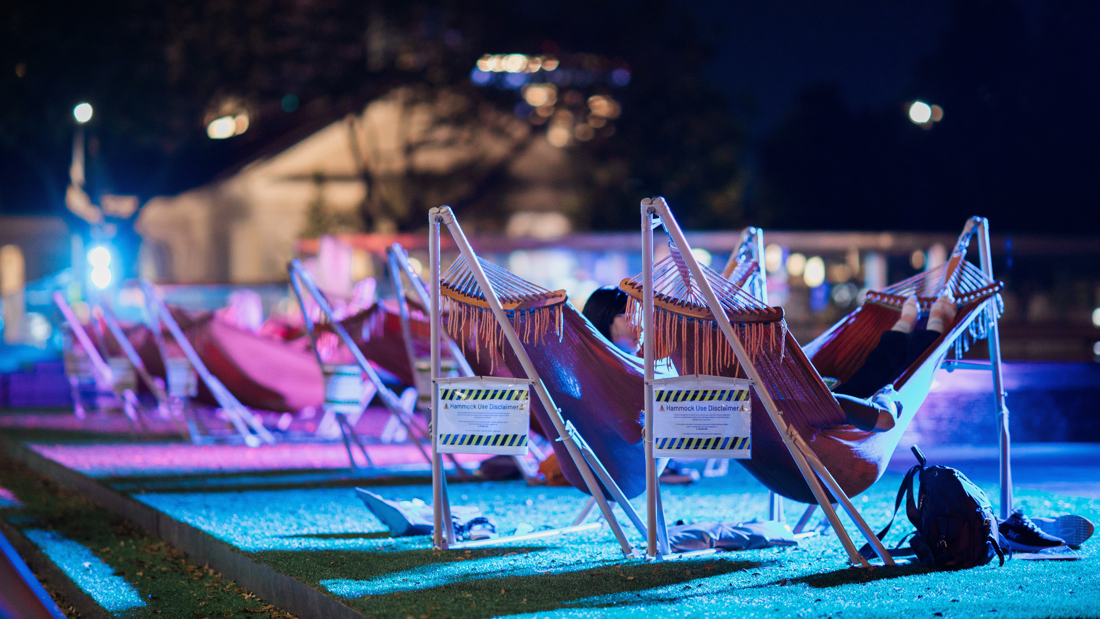A cinematic 16:9 unit still of a neon-lit public space in Singapore. A series of hammocks in repeating geometric patterns are bathed in high-contrast magenta and cyan light. A nocturnal study in urban leisure and atmospheric geometry, reflecting a modern cyber-noir aesthetic.