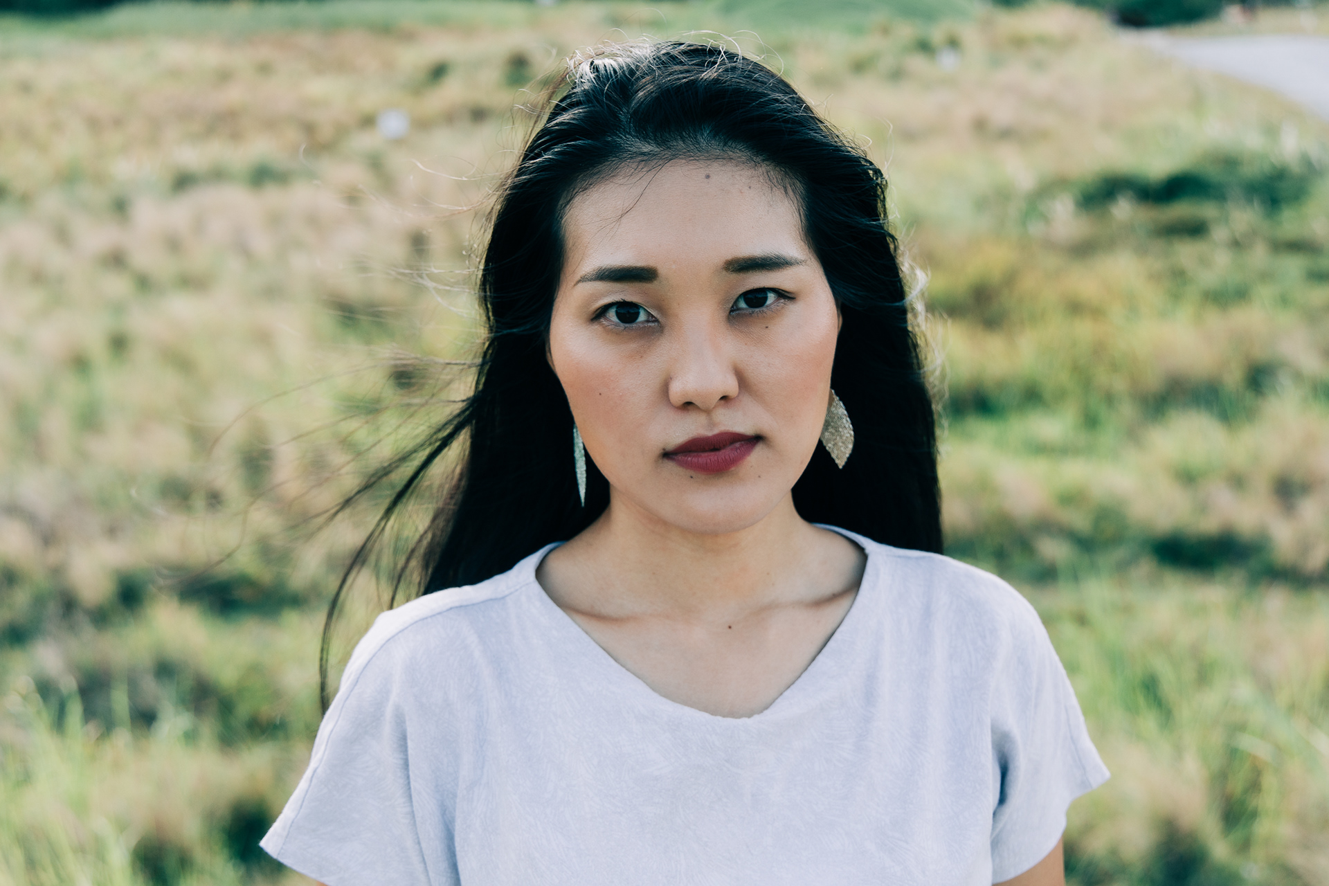 Cinematic high-fidelity headshot of a woman in an open field; 16:9 unit-still capturing a direct, candid gaze with soft atmospheric bloom and natural environmental textures.