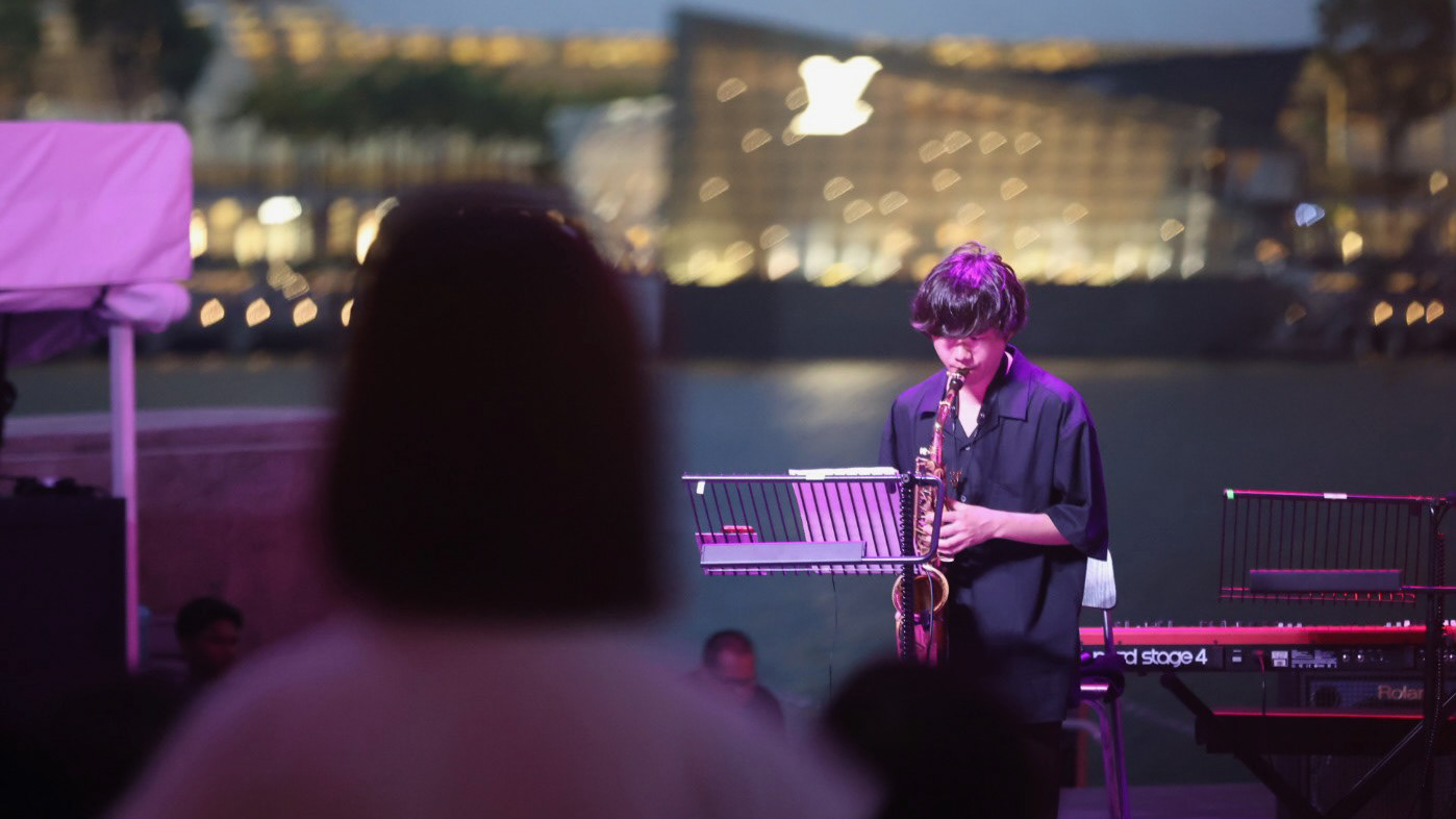 A 16:9 cinematic extraction of a saxophone player at the Esplanade Waterfront. The background features the blurred "atmospheric bloom" of the Marina Bay Sands and Louis Vuitton island store. Captured from the audience POV with a directorial focus on the interplay between the performer and the Singapore skyline.