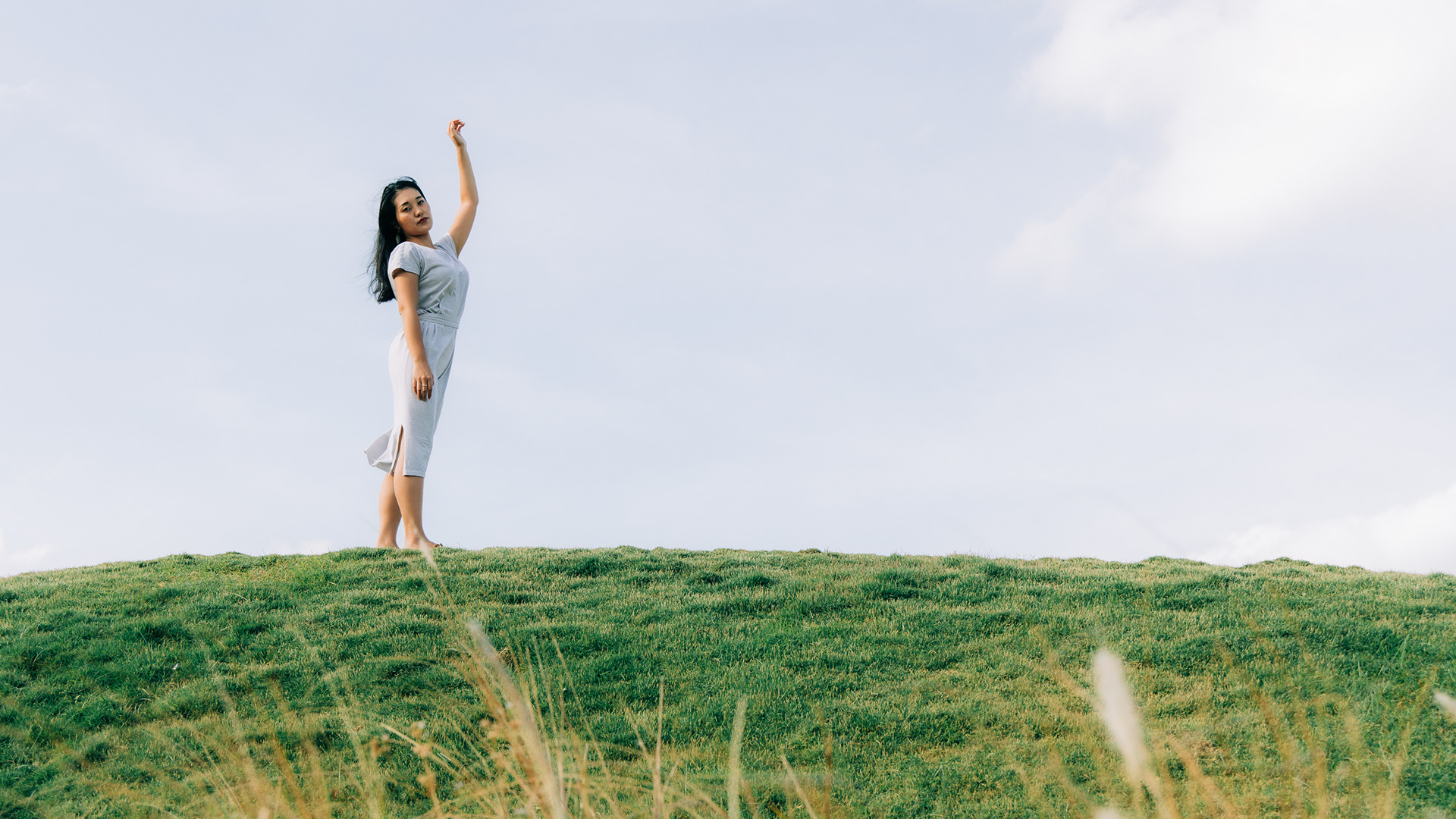 orizontal environmental portrait of a woman in a light-colored dress standing on a grassy hill against a vast, bright sky, reaching one arm upward.