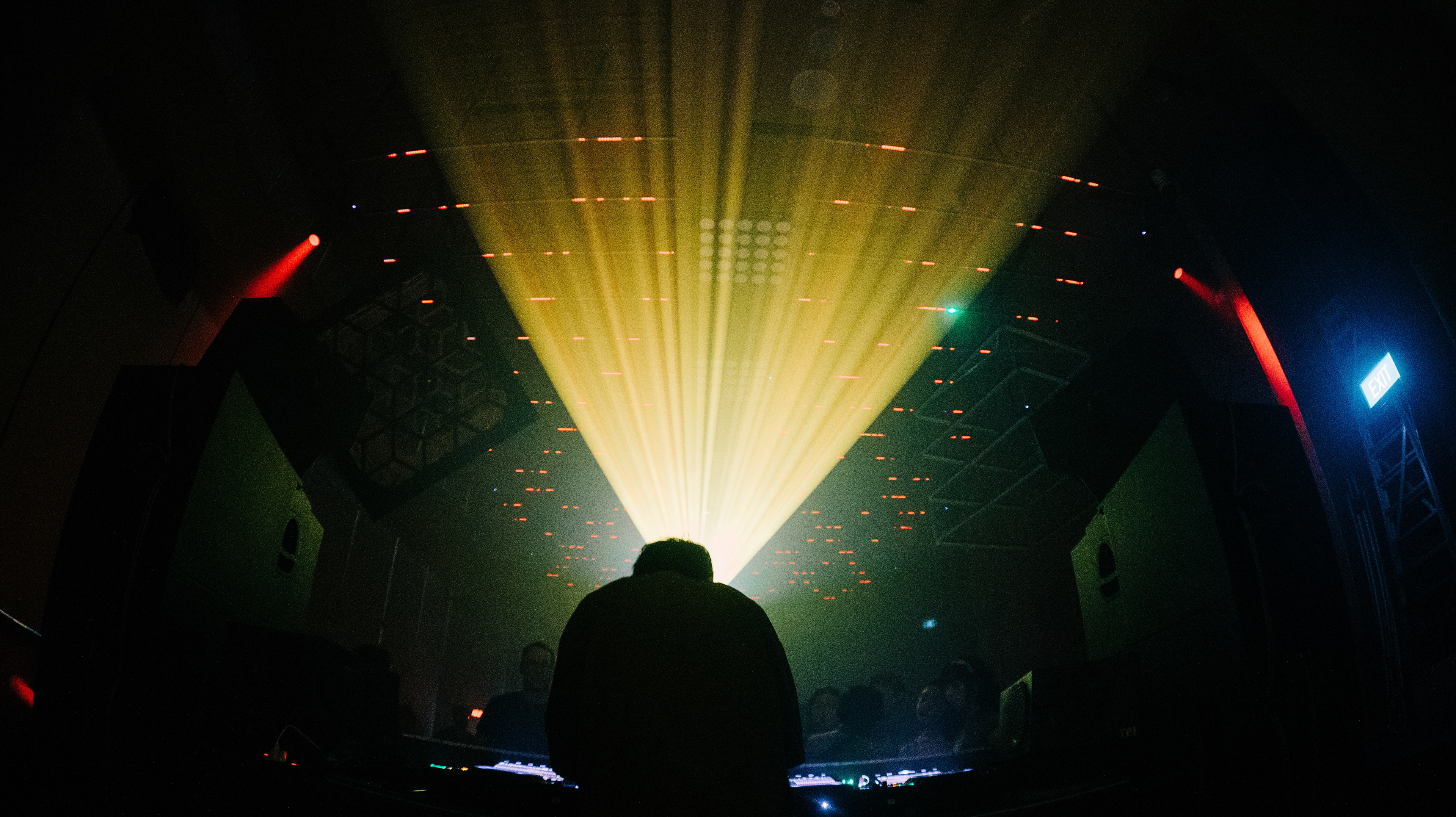A wide 16:9 cinematic extraction of a live music venue. High-intensity yellow light rays bisect the upper left frame, illuminating dust motes and atmospheric haze. Geometric, hexagonal ceiling installations create a heavy architectural silhouette against a concrete wall featuring the "Darker Than Wax" glowing logo. The crowd and DJ are captured from a voyeuristic, low-angle perspective, emphasizing the scale of the environment.