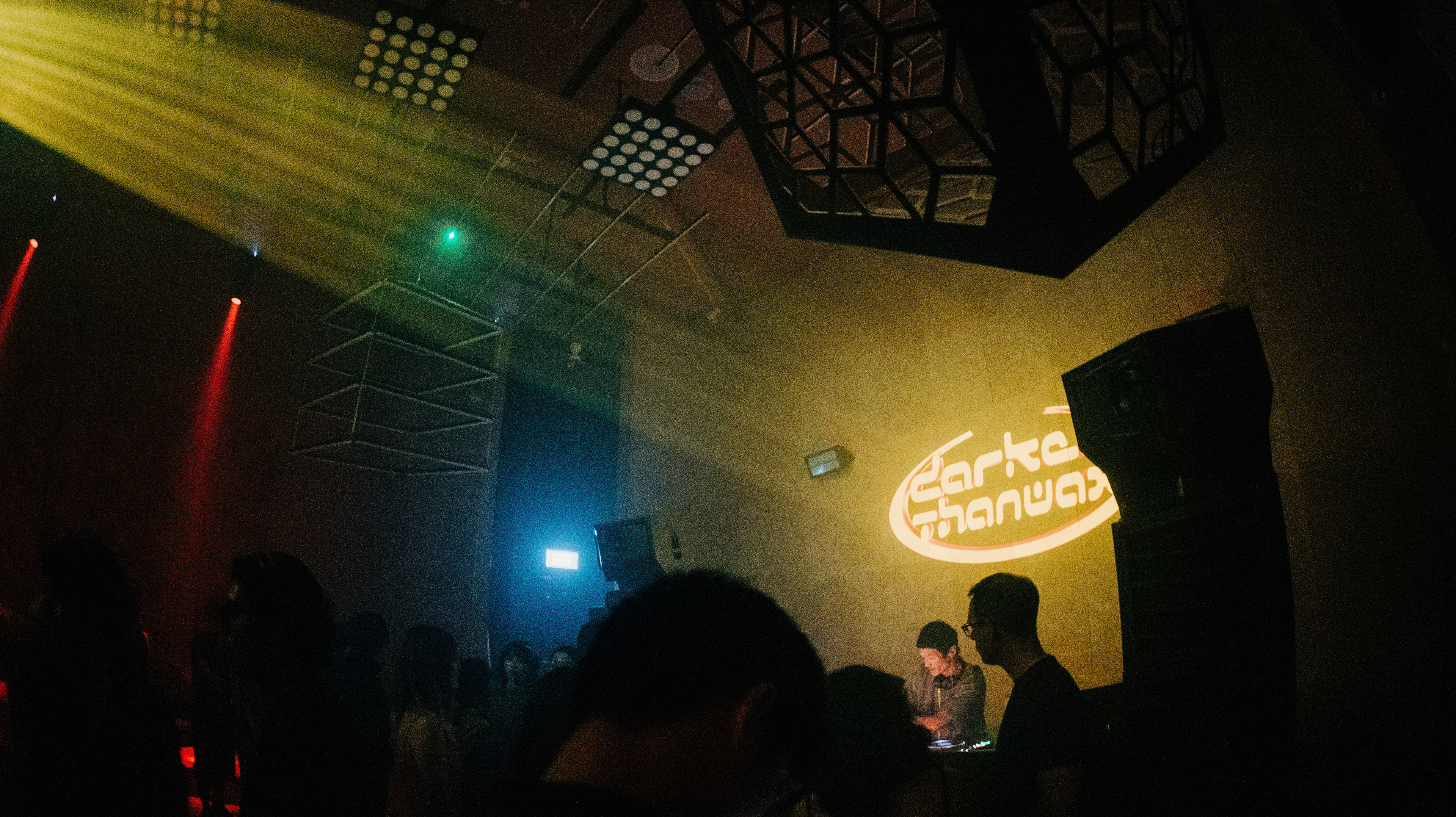 A wide 16:9 cinematic extraction of a live music venue. High-intensity yellow light rays bisect the upper left frame, illuminating dust motes and atmospheric haze. Geometric, hexagonal ceiling installations create a heavy architectural silhouette against a concrete wall featuring the "Darker Than Wax" glowing logo. The crowd and DJ are captured from a voyeuristic, low-angle perspective, emphasizing the scale of the environment.