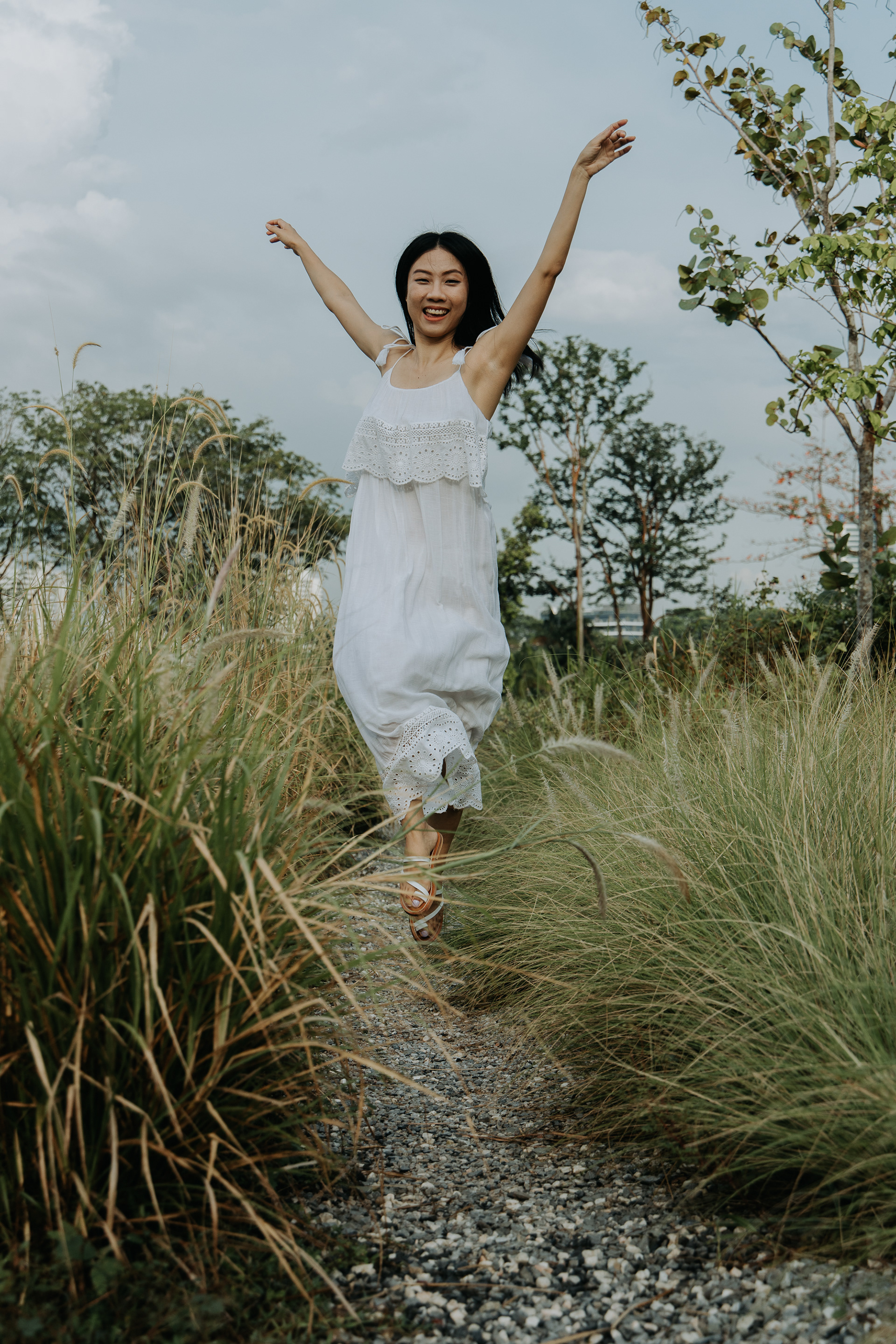 Vertical action portrait of a woman in a white dress running joyfully down a gravel path surrounded by tall grass in natural light.