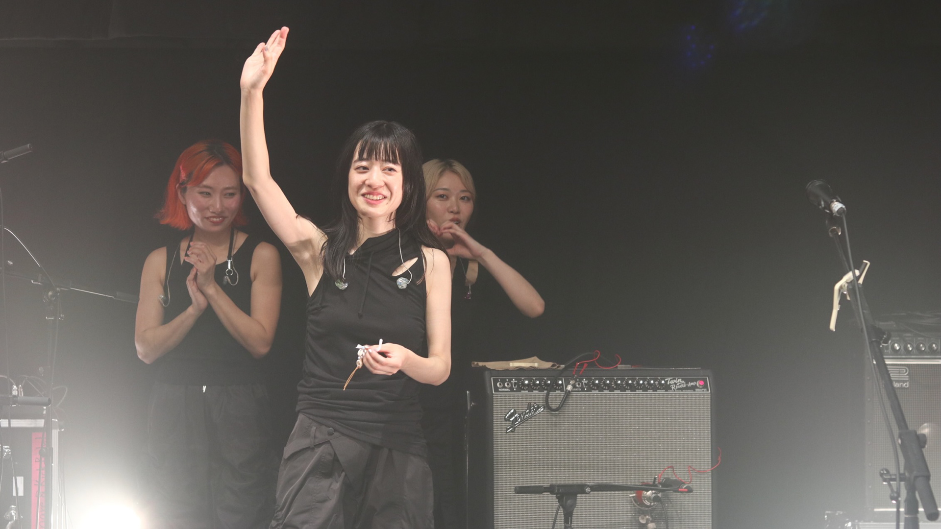 A documentary-style wide shot of Hitsujibungaku members Moeka Shiotsuka and Yurika Kasai waving to the crowd post-performance at the Esplanade. The composition balances the human connection of the artists with the technical backline, including their Fender amplifiers.