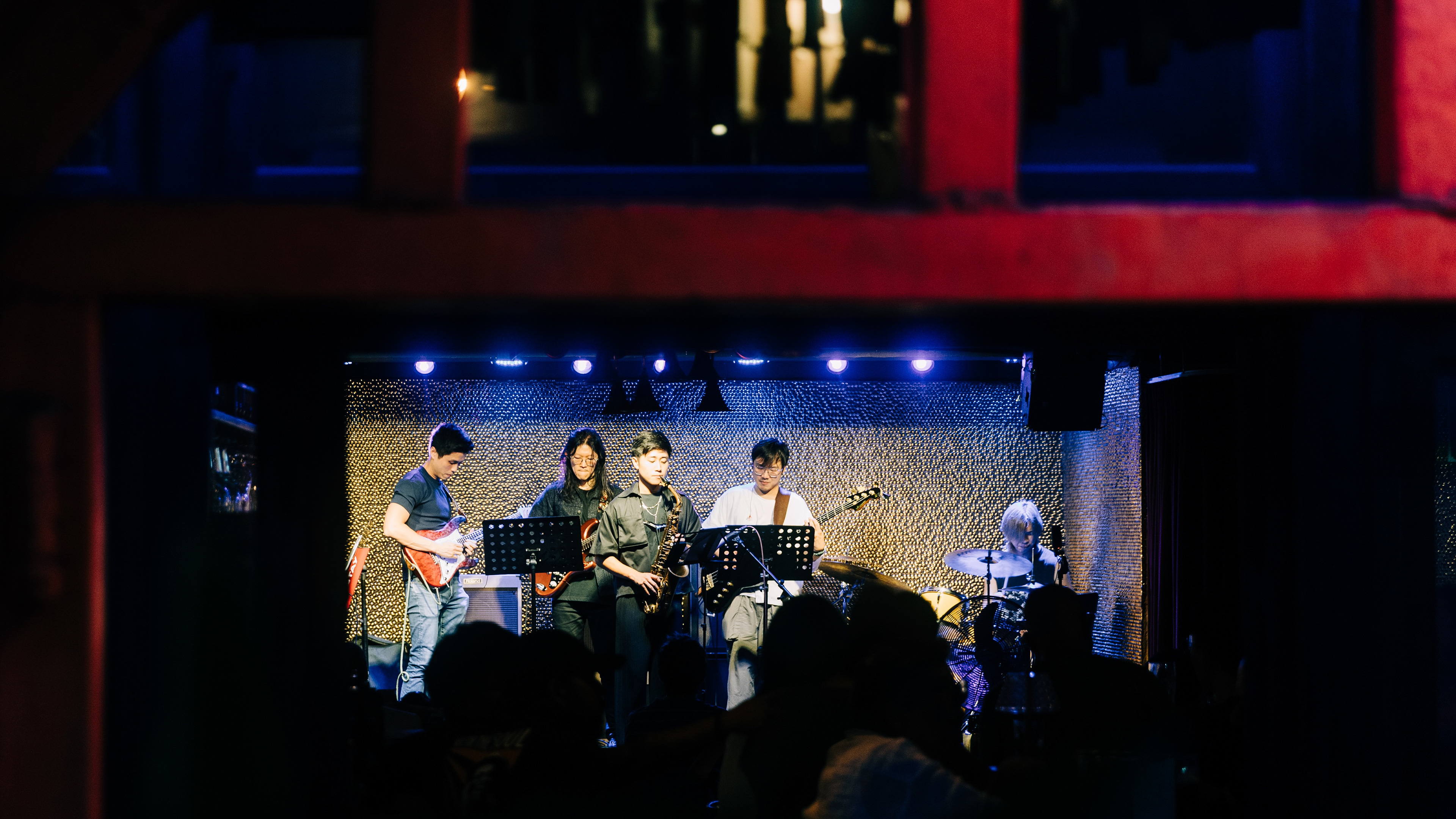 High-contrast cinematic wide shot of a jazz band performing on a stage with red and yellow lighting, framed through a structural architectural element in the foreground.
