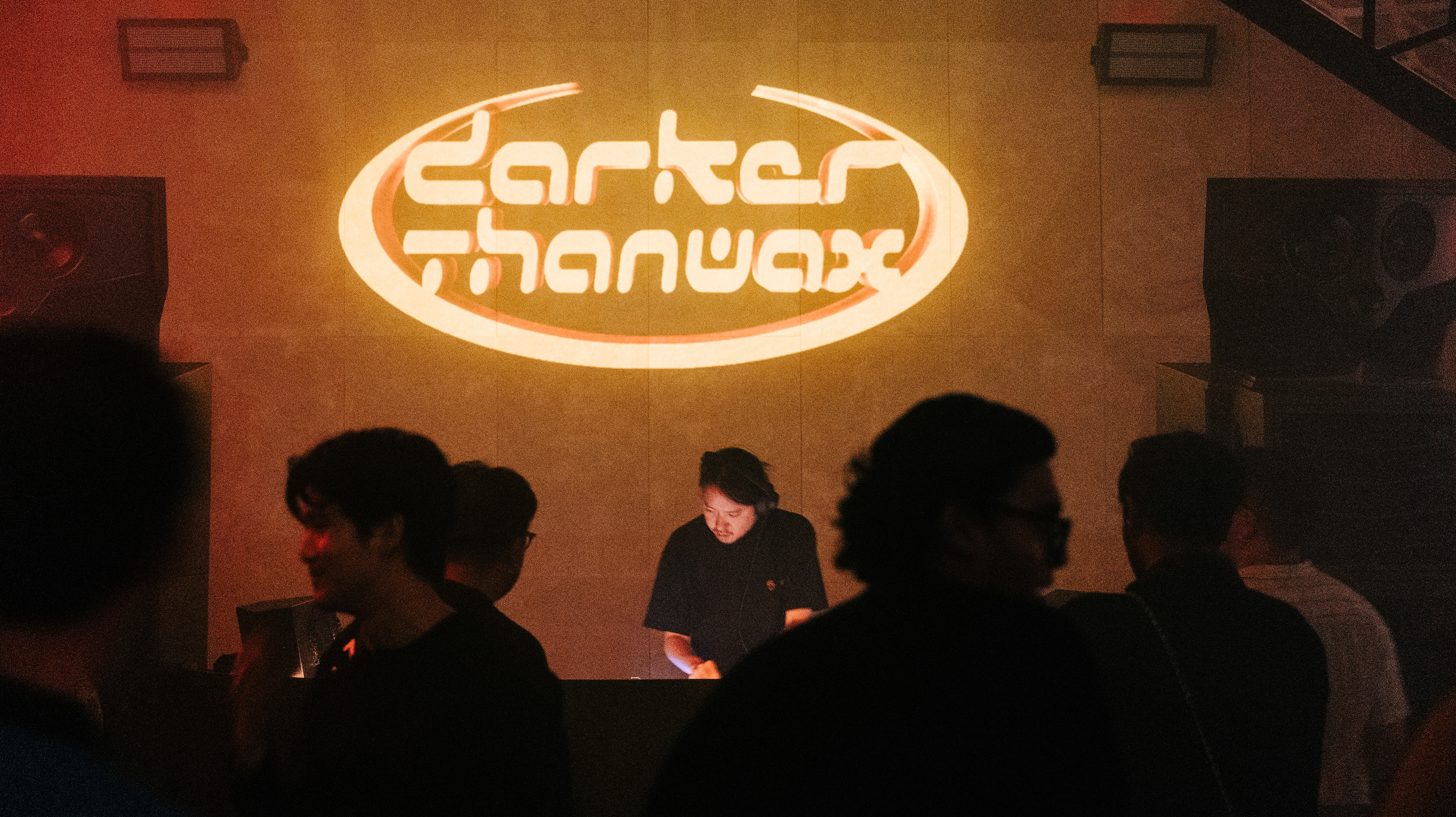 A medium-range cinematic shot within a 16:9 frame. The focus is on a DJ operating a mixer, positioned directly beneath a large, glowing orange "Darker Than Wax" neon-style projection. Foreground silhouettes of the audience are out of focus, creating a voyeuristic depth. The image features heavy film grain and a warm, low-light color palette, emphasizing the texture of the venue's concrete surfaces and the intensity of the performance.