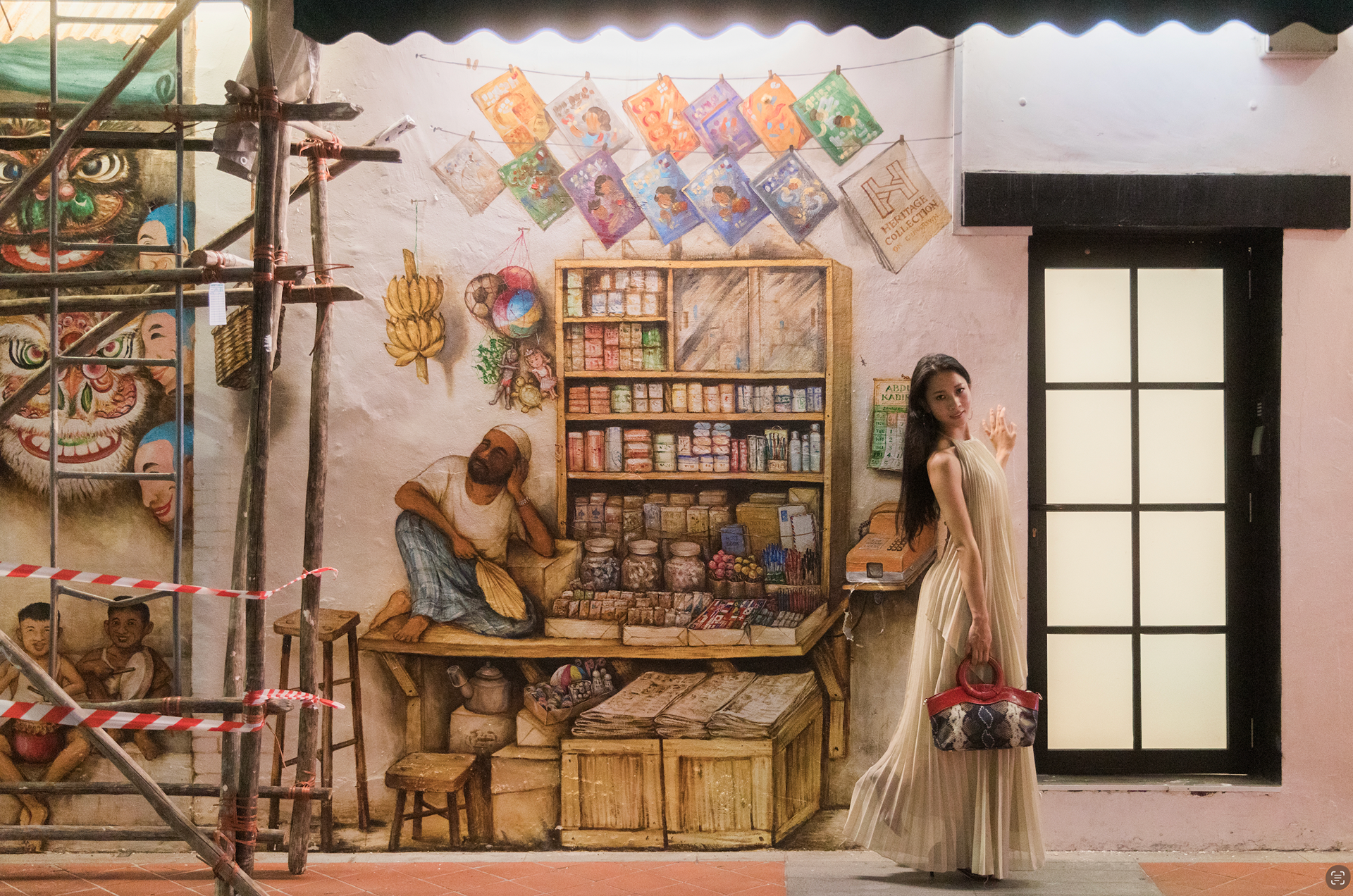 A detailed portrait study featuring a subject in traditional attire standing against a large-scale heritage mural wall in Singapore. The composition emphasizes light texture and cultural storytelling.