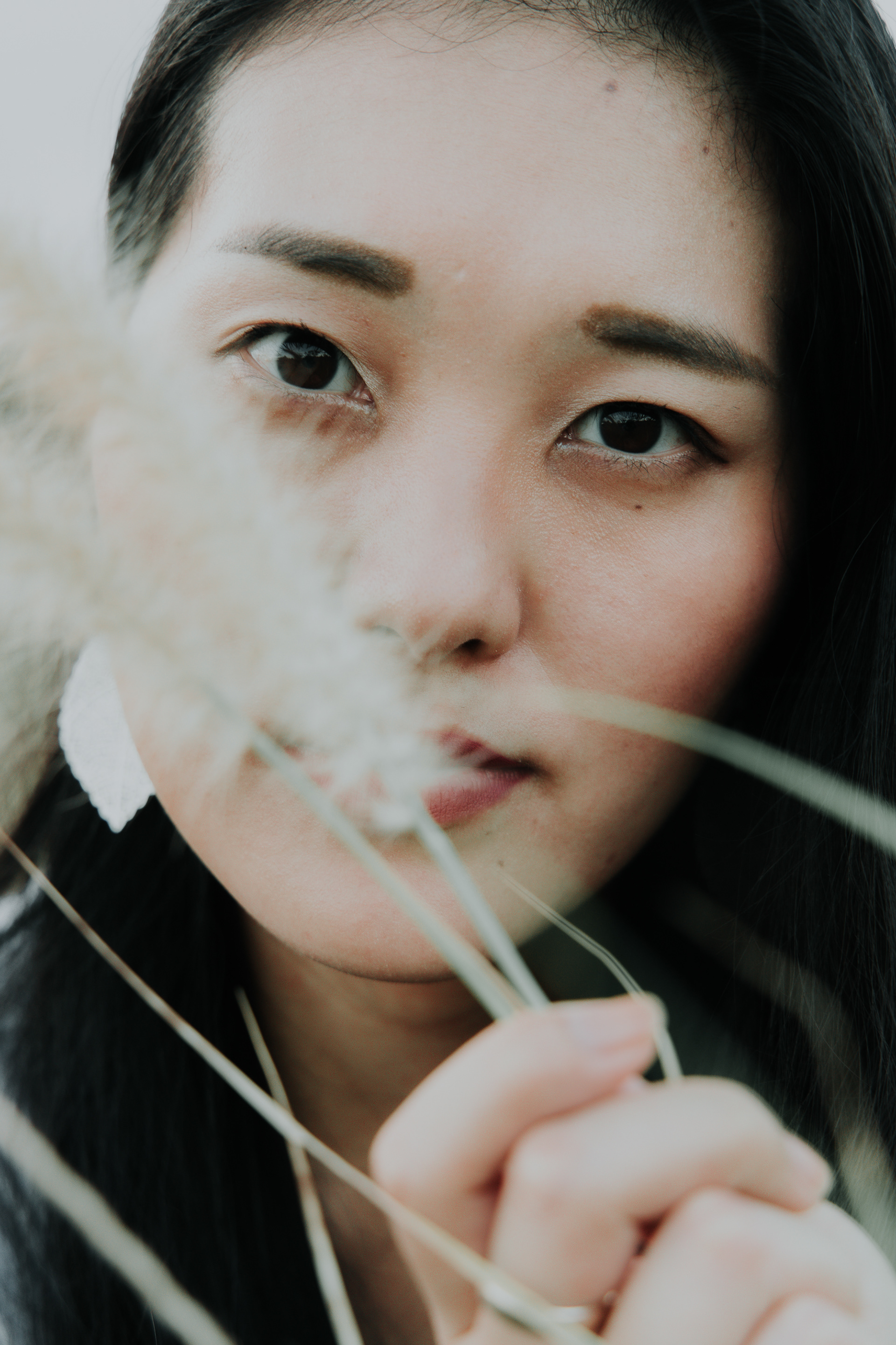 Vertical conceptual close-up portrait of a woman looking intensely at the camera, with the left side of her face obscured by tall grass, featuring sharp focus on one eye.
