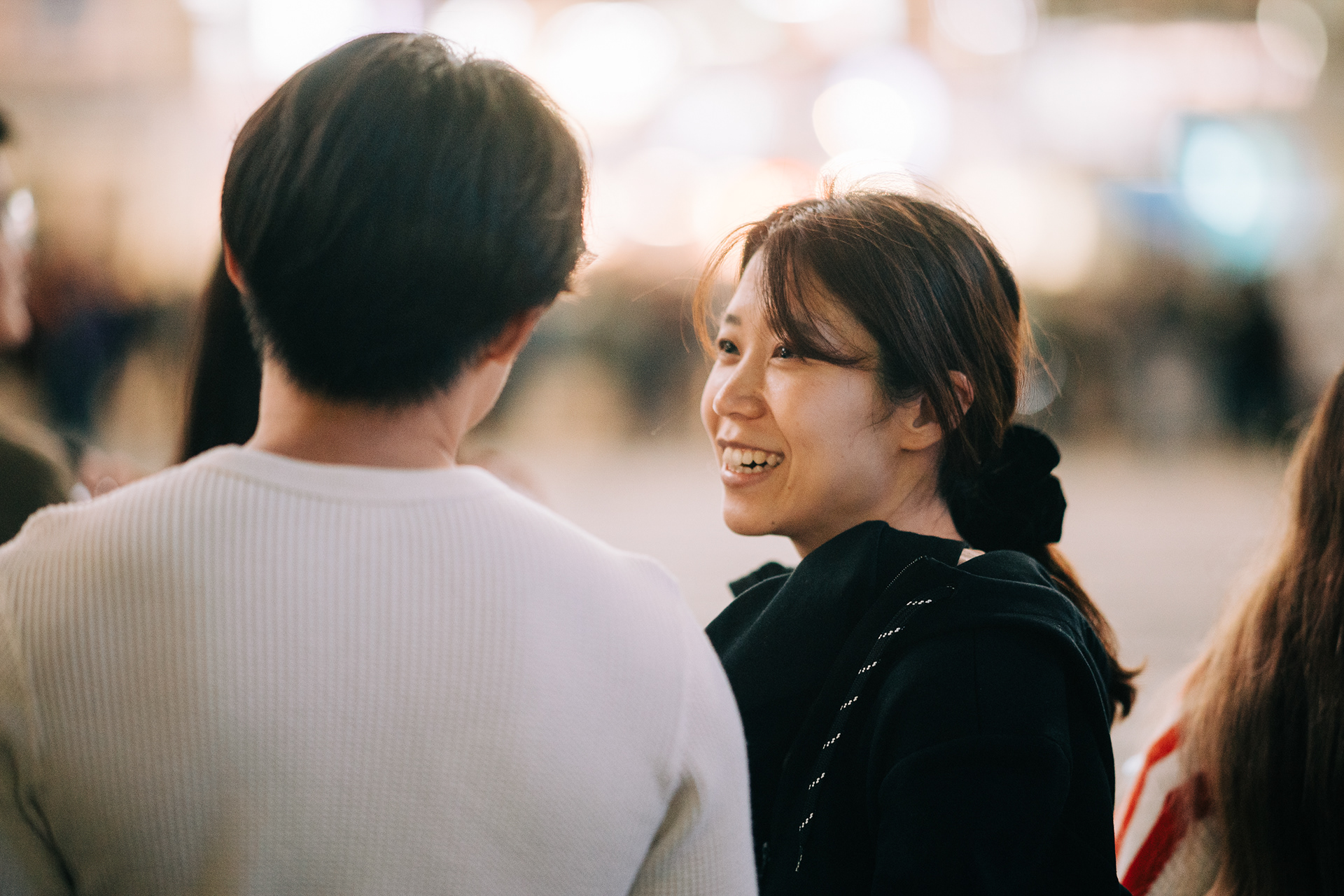 Candid over-the-shoulder street photography of a woman smiling during a conversation at night, with soft golden bokeh background.