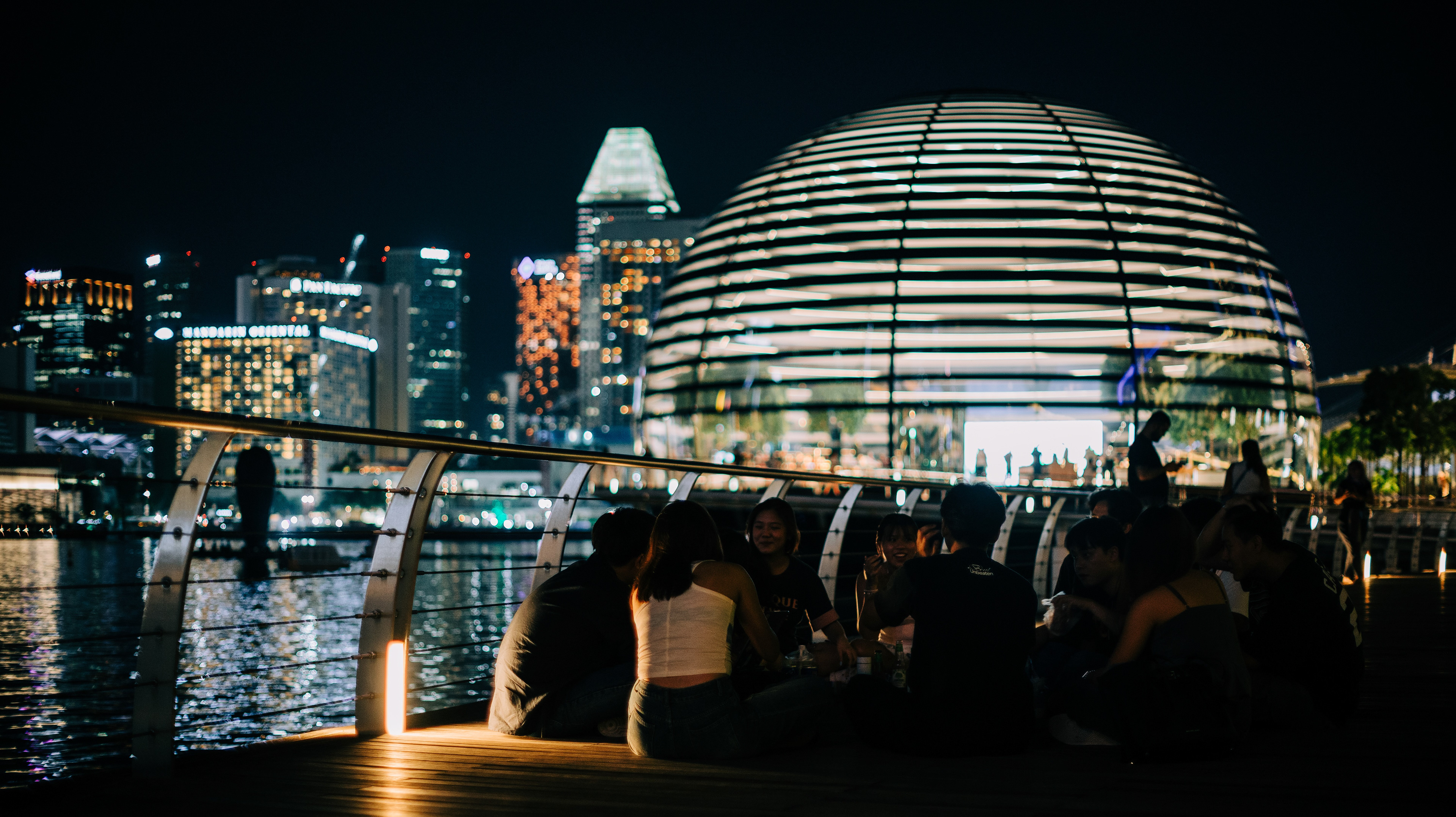 A wide-angle cinematic photograph of a group of people sitting on the Marina Bay Sands boardwalk at night. The illuminated Apple Marina Bay Sands dome and the Singapore city skyline glow in the background, illustrating the intersection of human connection and grand architecture.