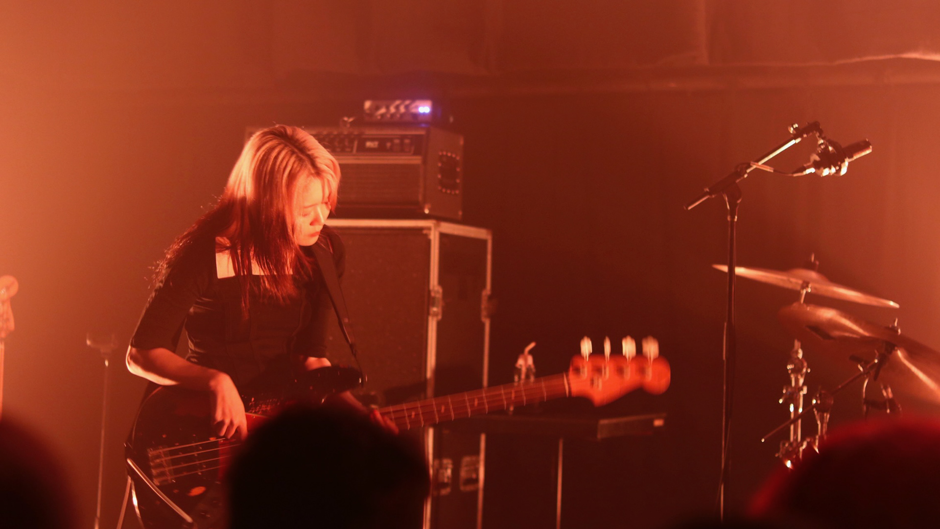 A cinematic 16:9 archival extraction of Hitsujibungaku bassist Yurika Kasai. She is captured in a deliberately structured, neutral pose under monochromatic amber lighting, emphasizing her focused mastery. The background features the "mechanical soul" of the stage architecture and amplifier stacks.
