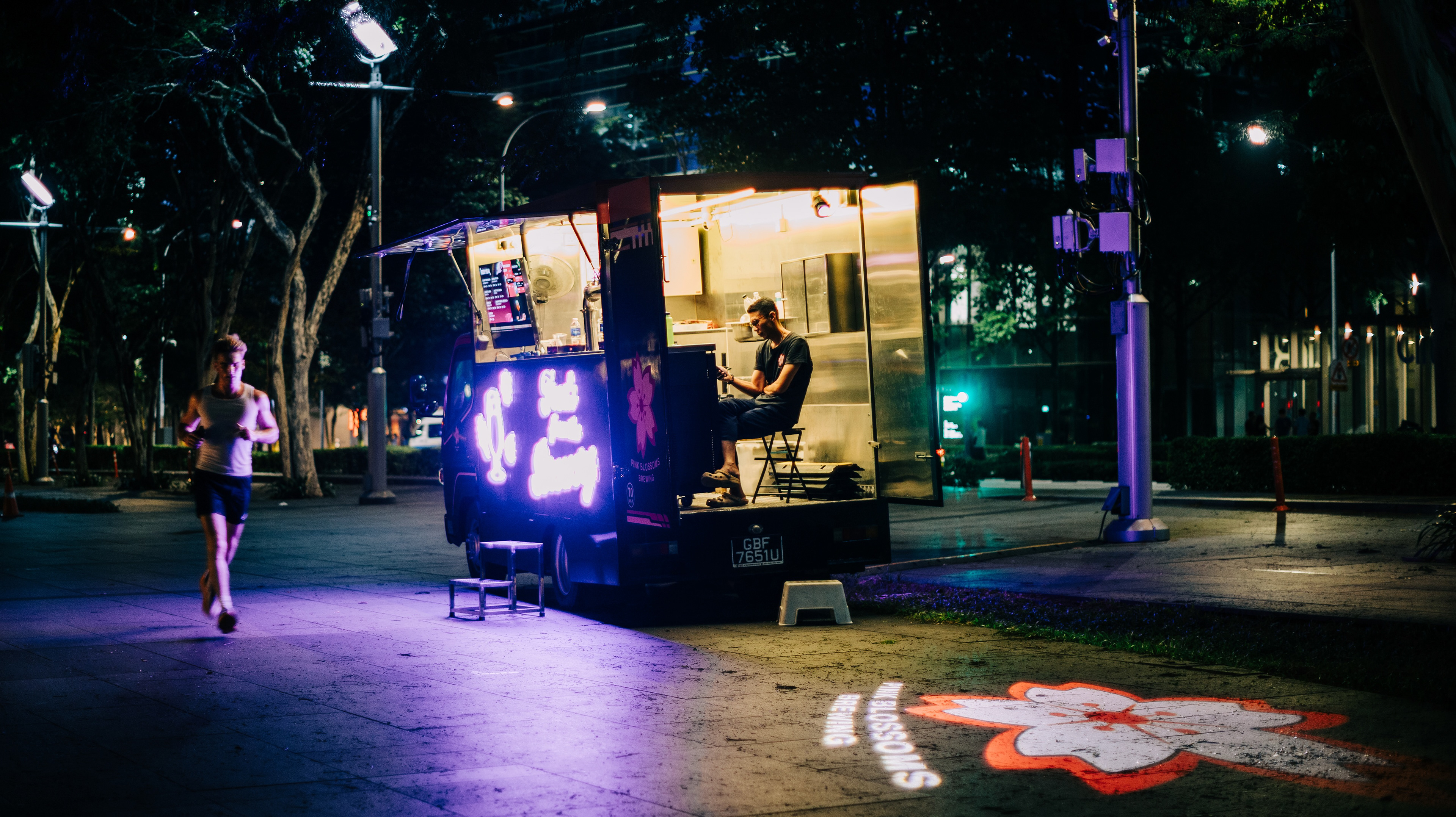 A cinematic night-time street photograph of a mobile kiosk illuminated by purple neon signs. A runner passes by in a motion blur while a lone vendor sits inside, showcasing the high-contrast shadows and atmospheric lighting of a Singapore city night.
