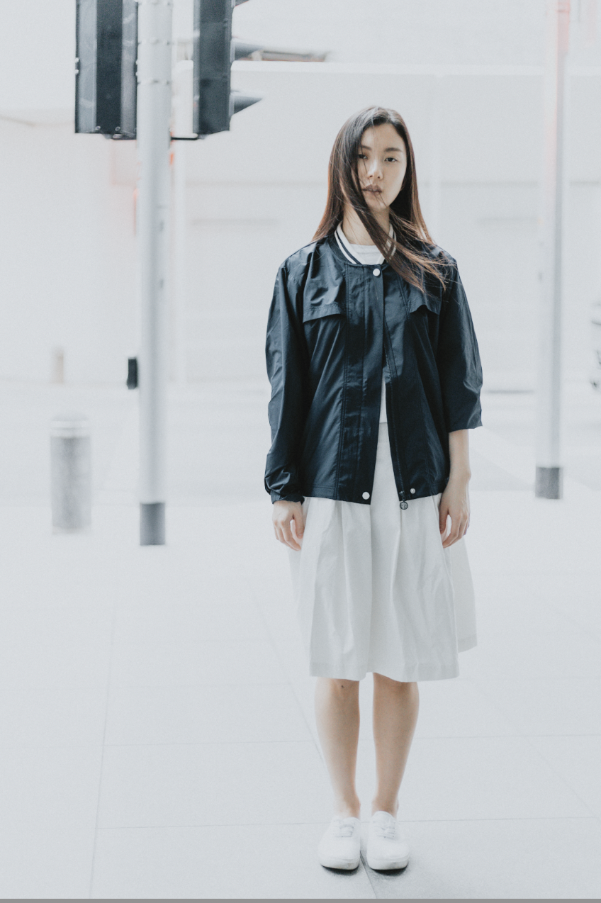 Vertical full-body urban portrait of a woman in a black jacket and white skirt standing in a minimalist, bright city environment.