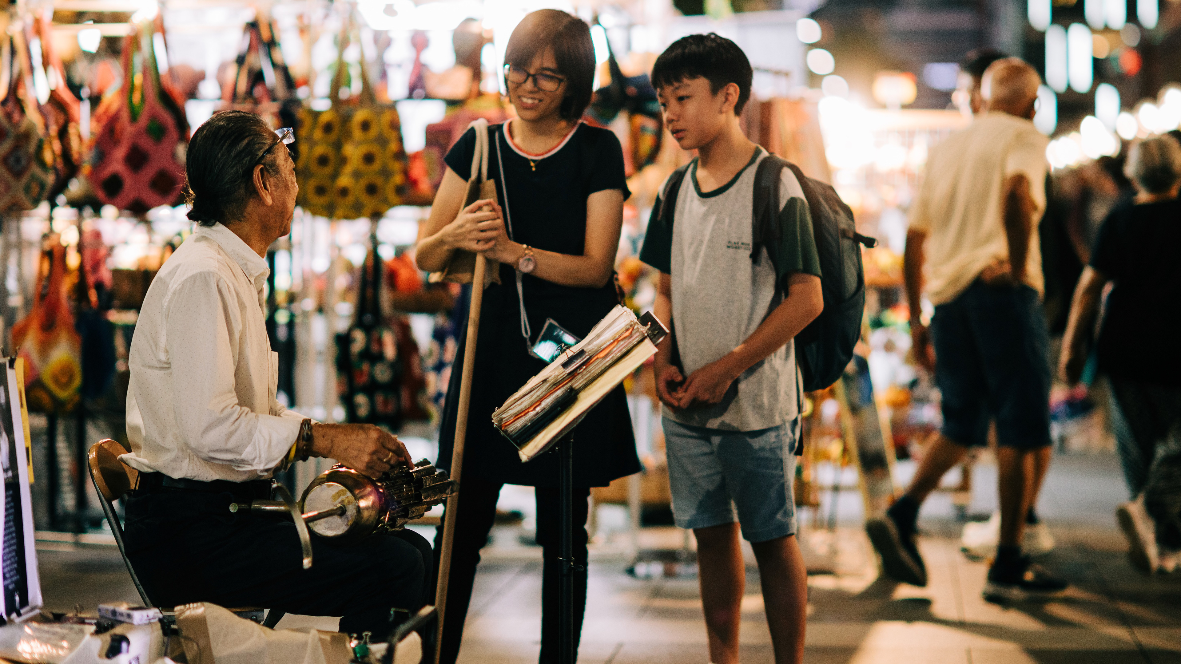 A 16:9 cinematic unit still capturing a traditional street musician performing for a mother and son in Singapore. High-fidelity documentary style with a focus on human connection, cultural heritage, and the soft nocturnal atmosphere of a local night market.