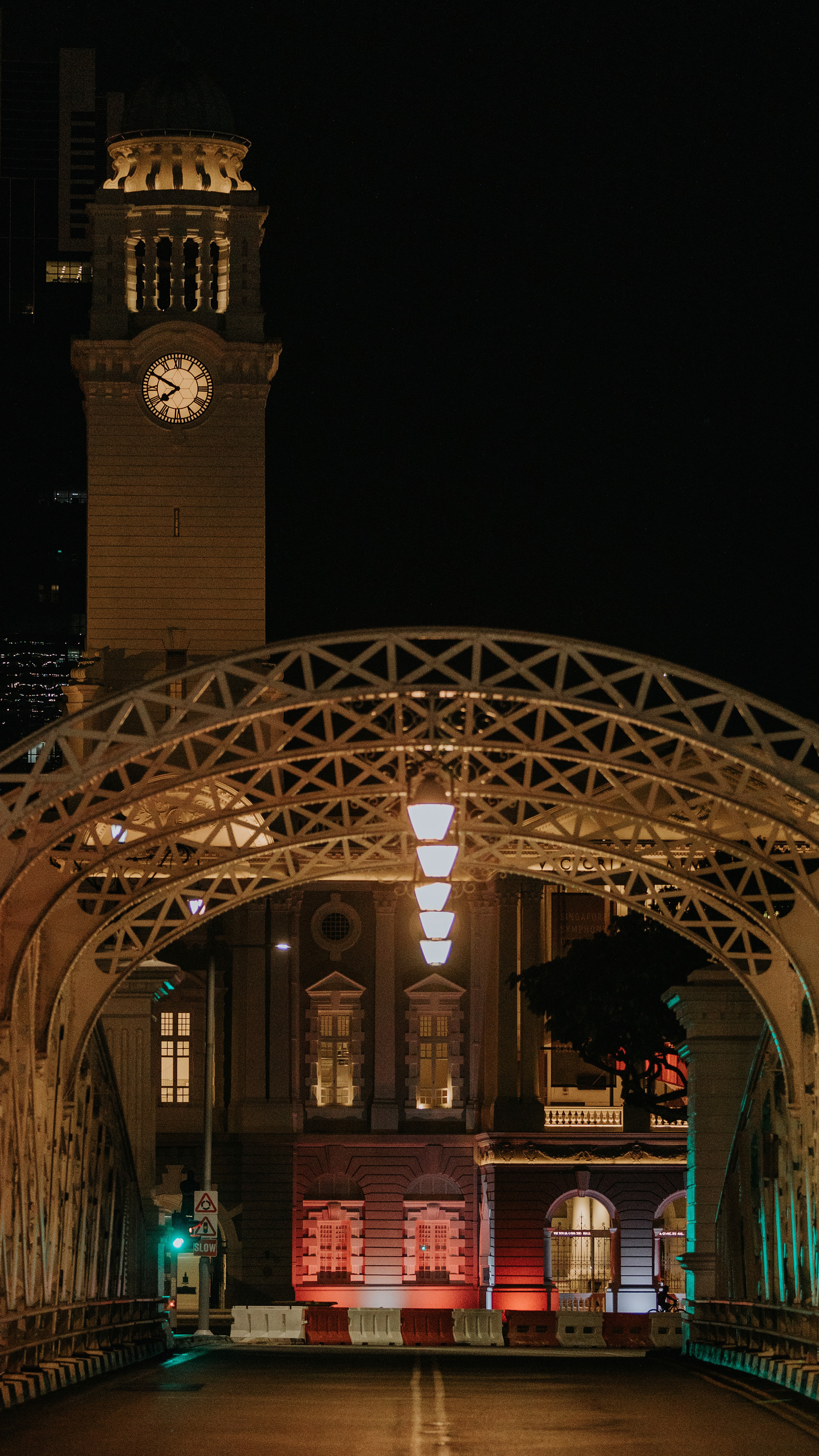 A high-fidelity vertical unit still capturing the neoclassical architecture of the Victoria Theatre and Concert Hall, framed symmetrically by the historical steel arches of the Anderson Bridge in Singapore. A nocturnal study in geometric lines, leading composition, and the 'Mechanical Soul' of the urban structure.