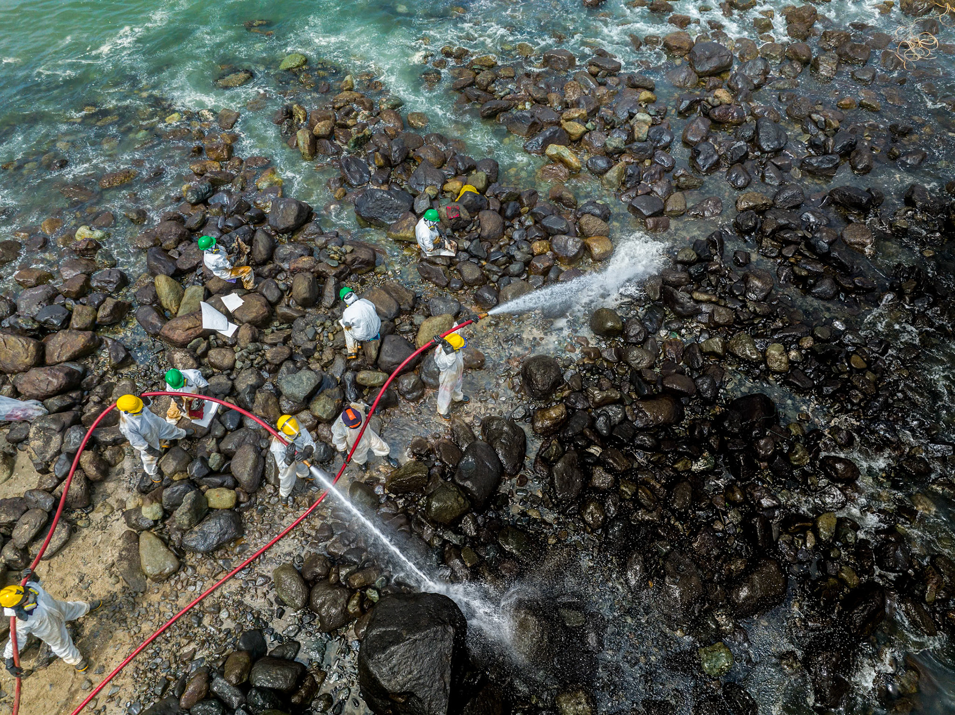 Aerial view of cleanup efforts after oil spill at "La Pampilla" refinery, Ancon, Lima, Peru on 2022. Workers in protective suits cleaning rocky shoreline.