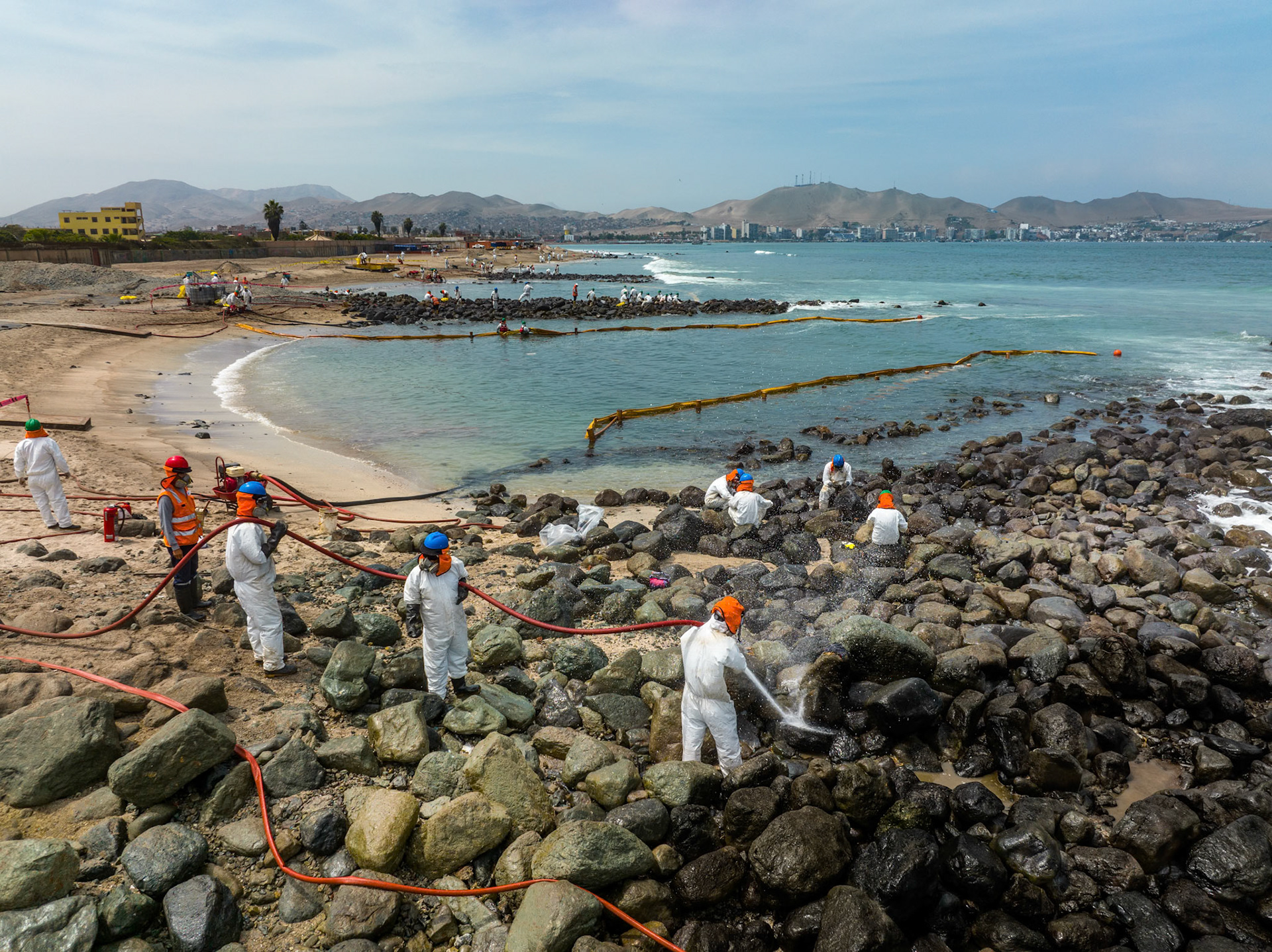 Aerial view of cleanup efforts after oil spill at "La Pampilla" refinery, Ancon, Lima, Peru on 2022. Workers in protective suits cleaning rocky shoreline.