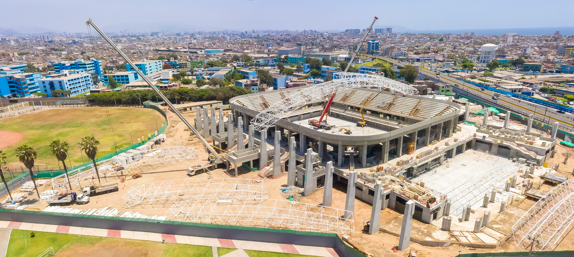 Aerial image of the construction of a coliseum, showing the gray work, the structures, the work of the cranes, metal structures of the roofs and very small people. Captured with a drone, under the light of a sunny day, in the city of Lima, Peru.