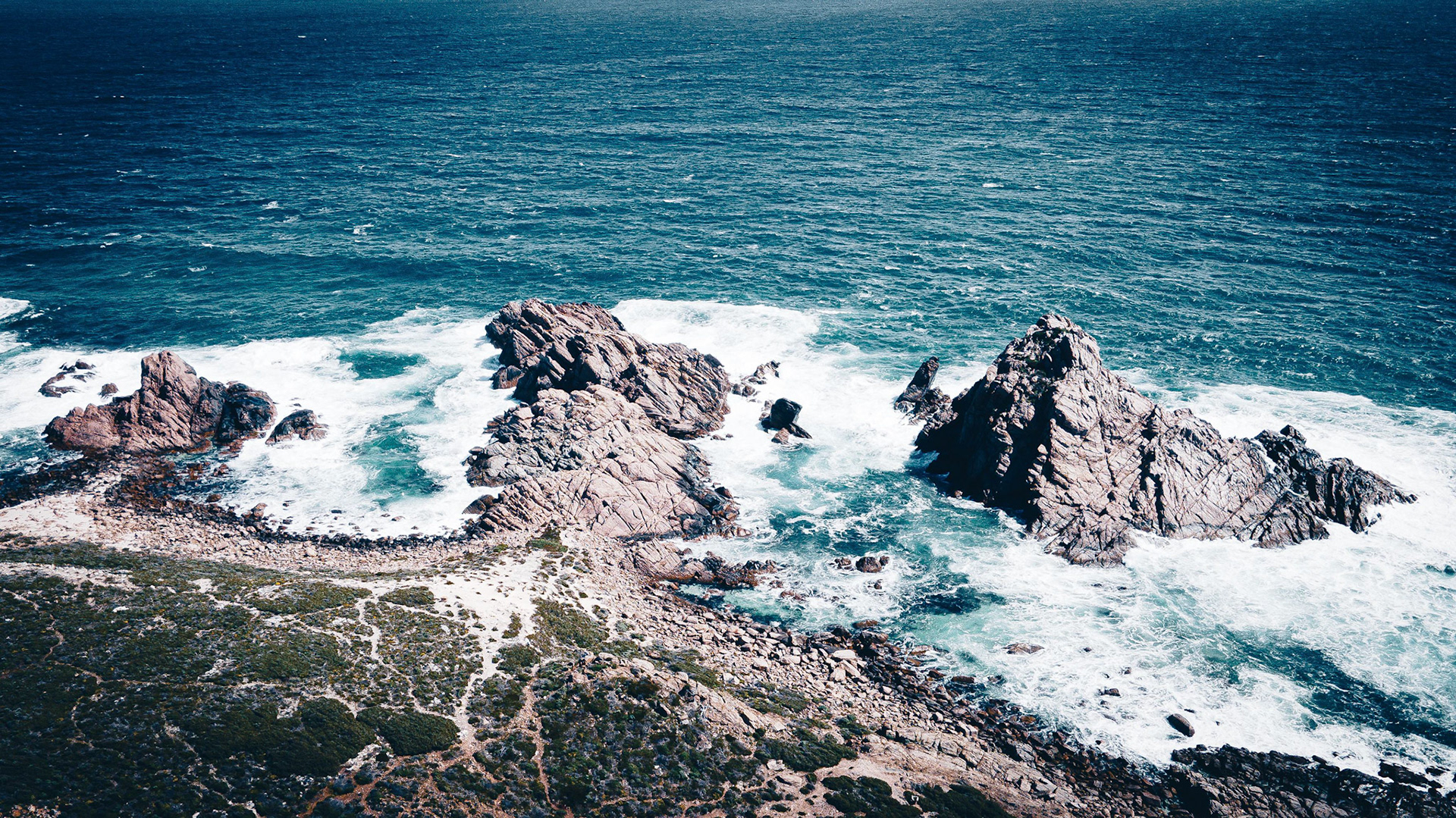 A stunning aerial view captures the rugged coastal landscape with dramatic rock formations surrounded by the vibrant blue ocean. The waves crash against the rocks, creating a dynamic contrast between the calm sea and the rough terrain. The lush greenery on the shoreline adds a touch of natural beauty, enhancing the scene's overall appeal.