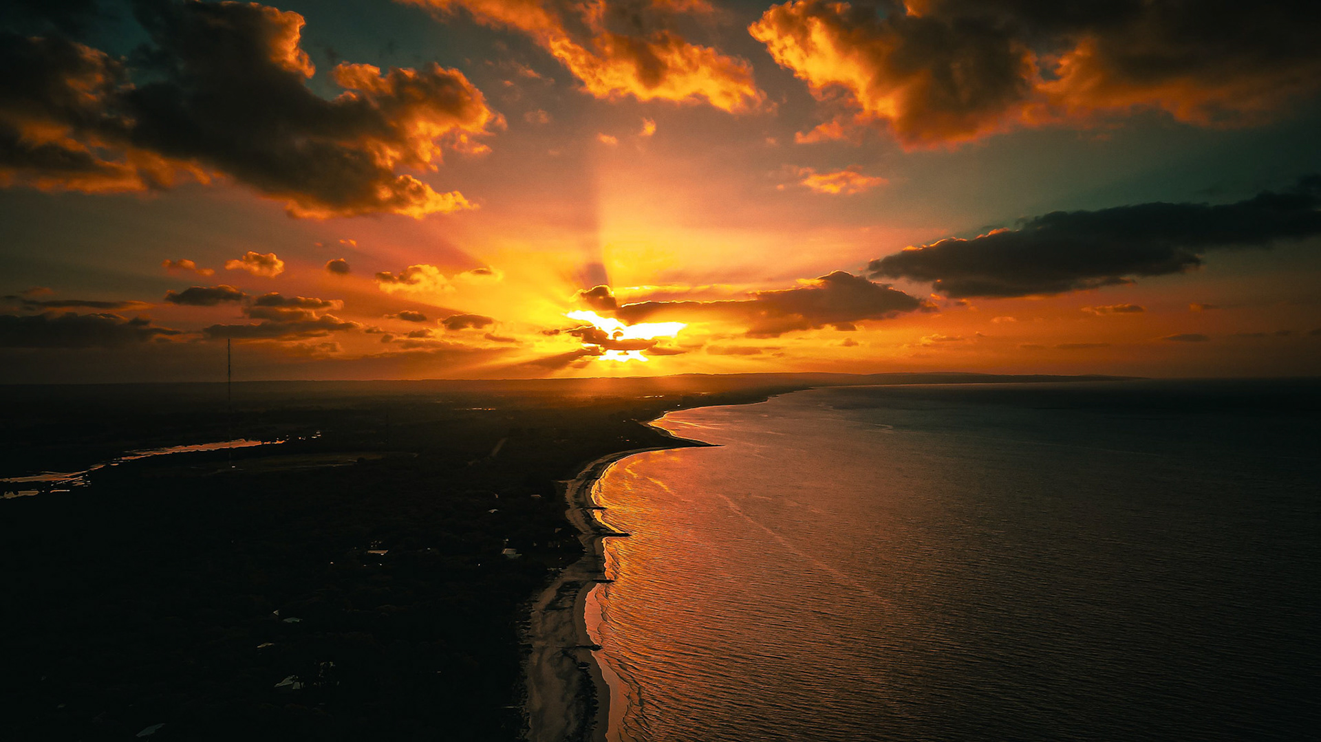A breathtaking aerial view showcasing a stunning sunset casting vibrant hues over the ocean and coastline. The clouds are illuminated with golden and orange tones as the sun dips below the horizon, creating a serene and dramatic scene. The shoreline curves gently, framing the tranquil waters that reflect the sky's warm colors.
