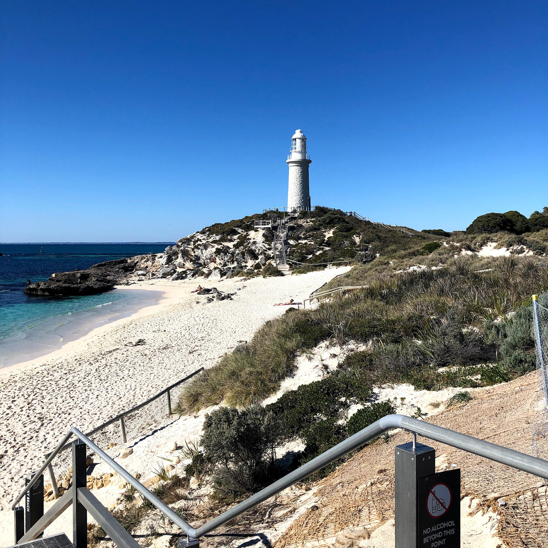 A white lighthouse stands on a grassy hill, overlooking a sandy beach and clear ocean. The bright blue sky enhances the serene atmosphere.