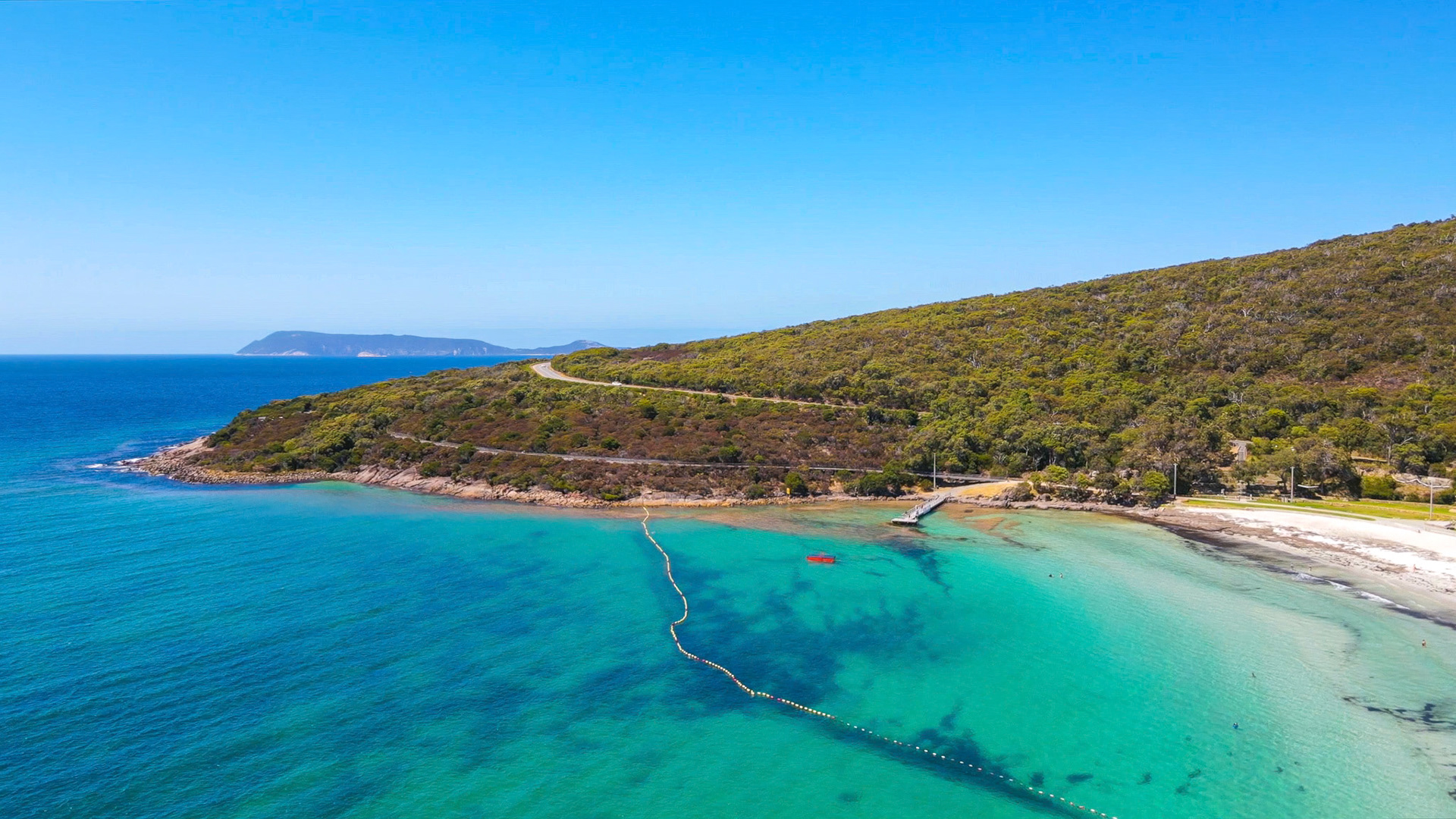 An aerial view showcases a serene coastal landscape with turquoise waters, sandy beach, and a winding road along the lush hillside.