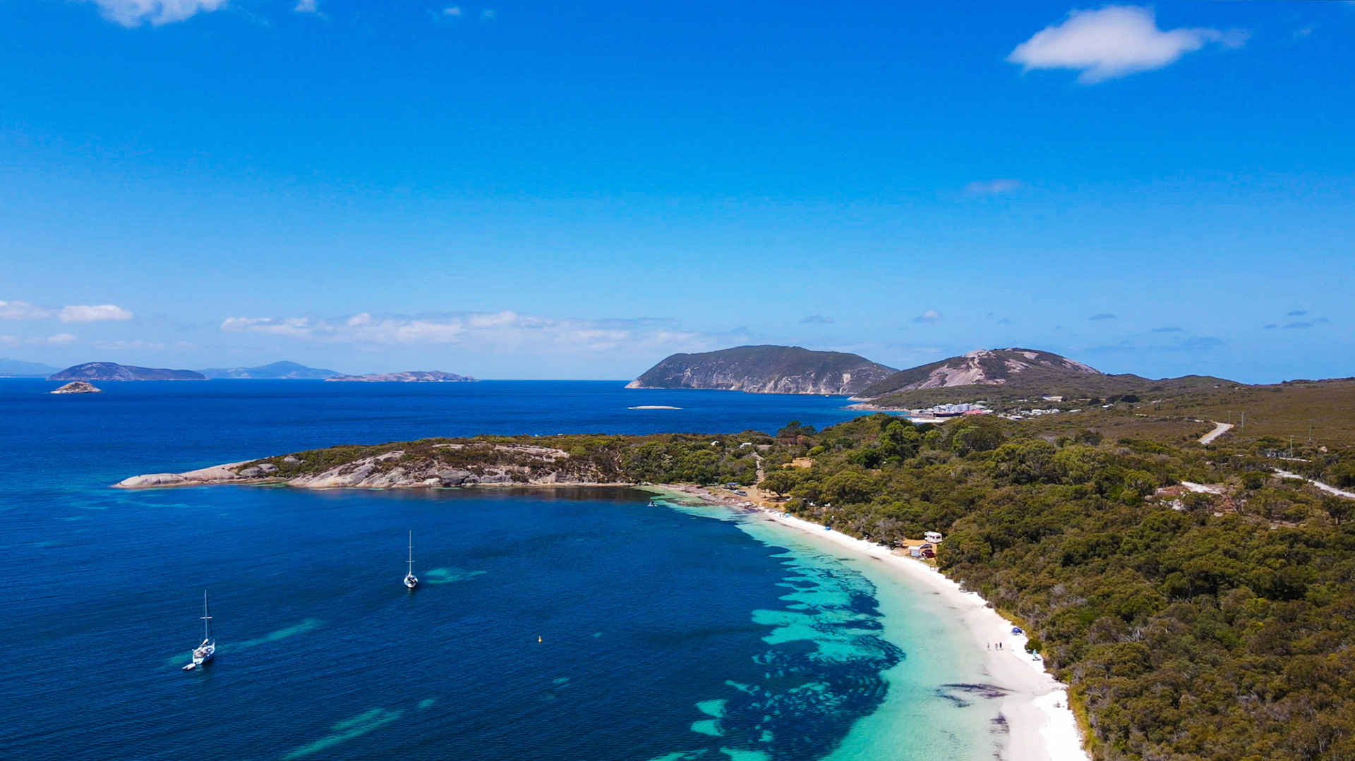 An aerial view showcases a serene beach and ocean landscape. Sailboats rest in tranquil waters, surrounded by lush greenery.