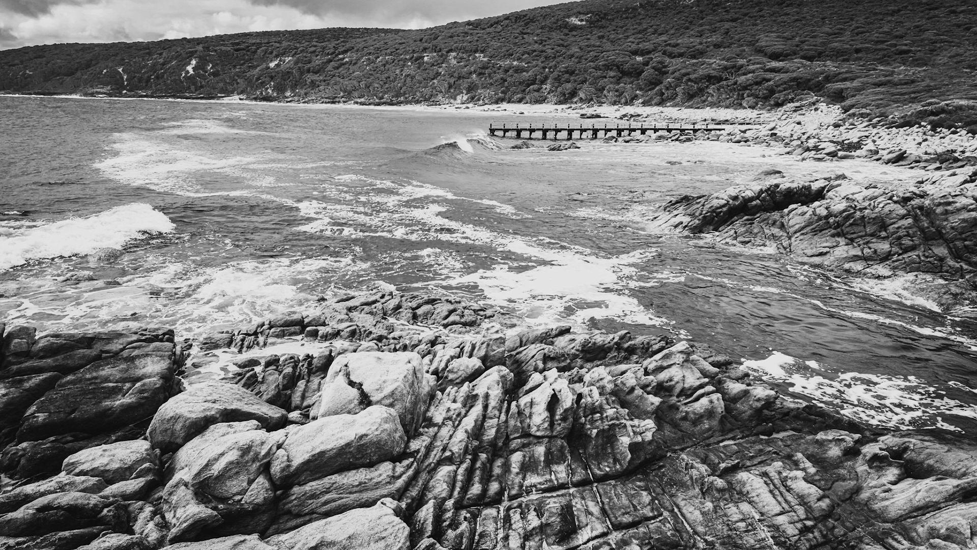 A stunning black-and-white image captures a rugged coastal rock formation meeting the ocean's waves under a cloudy sky. The texture of the rocks is highlighted, creating a sense of drama and timeless natural beauty. Waves crash against the shore, and a small wooden pier extends into the water, adding a human element to the otherwise untamed landscape.