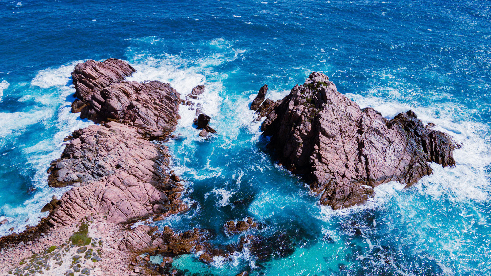 An aerial photograph capturing the stunning contrast of a rocky coastline against the vibrant blue hues of the ocean. The rough texture of the rocks complements the restless waves, showcasing nature's raw beauty and dynamic forces at work. Ideal for use in travel, nature, and coastal tourism projects.