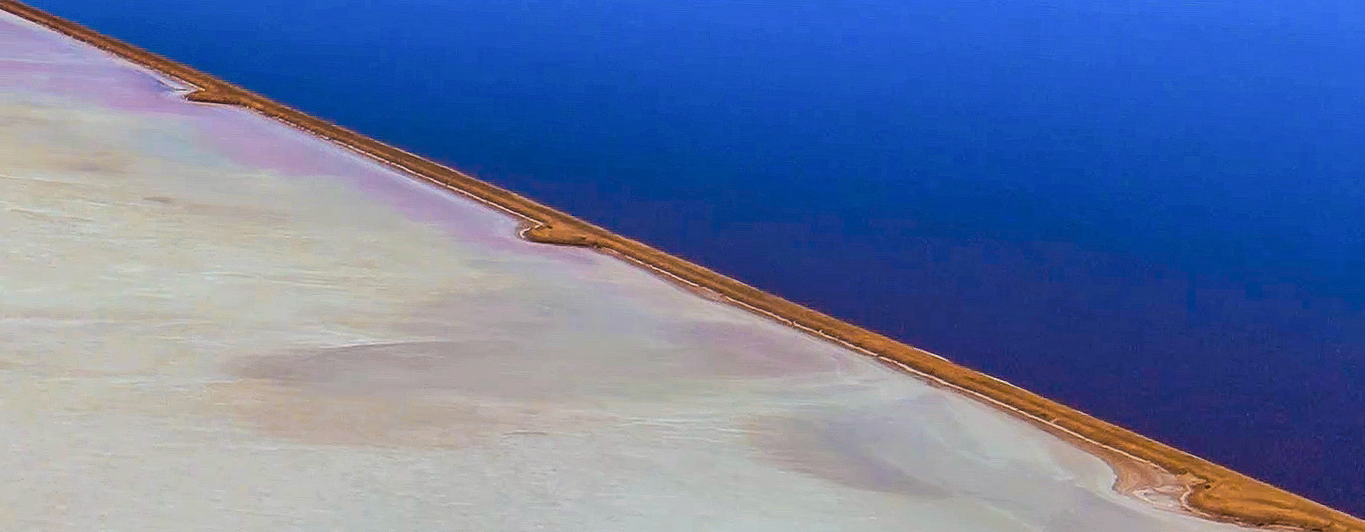 An aerial view highlights the contrast between a salt flat and deep blue water, showcasing unique geological textures and colors.