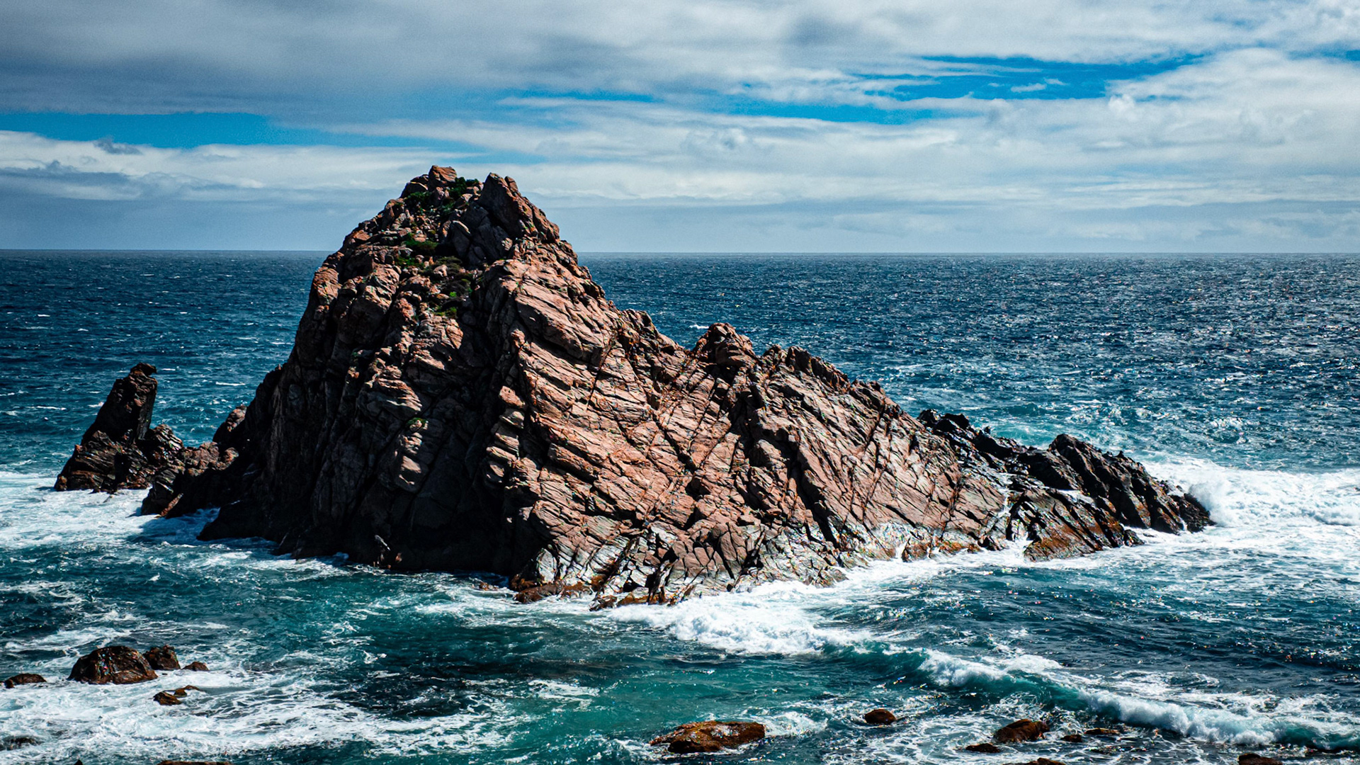 Stunning view of a rugged rocky outcrop surrounded by the crashing waves of a deep blue ocean under a partly cloudy sky. The dramatic formation showcases nature's raw beauty, making it an ideal scene for depicting serenity, inspiration, and natural power.