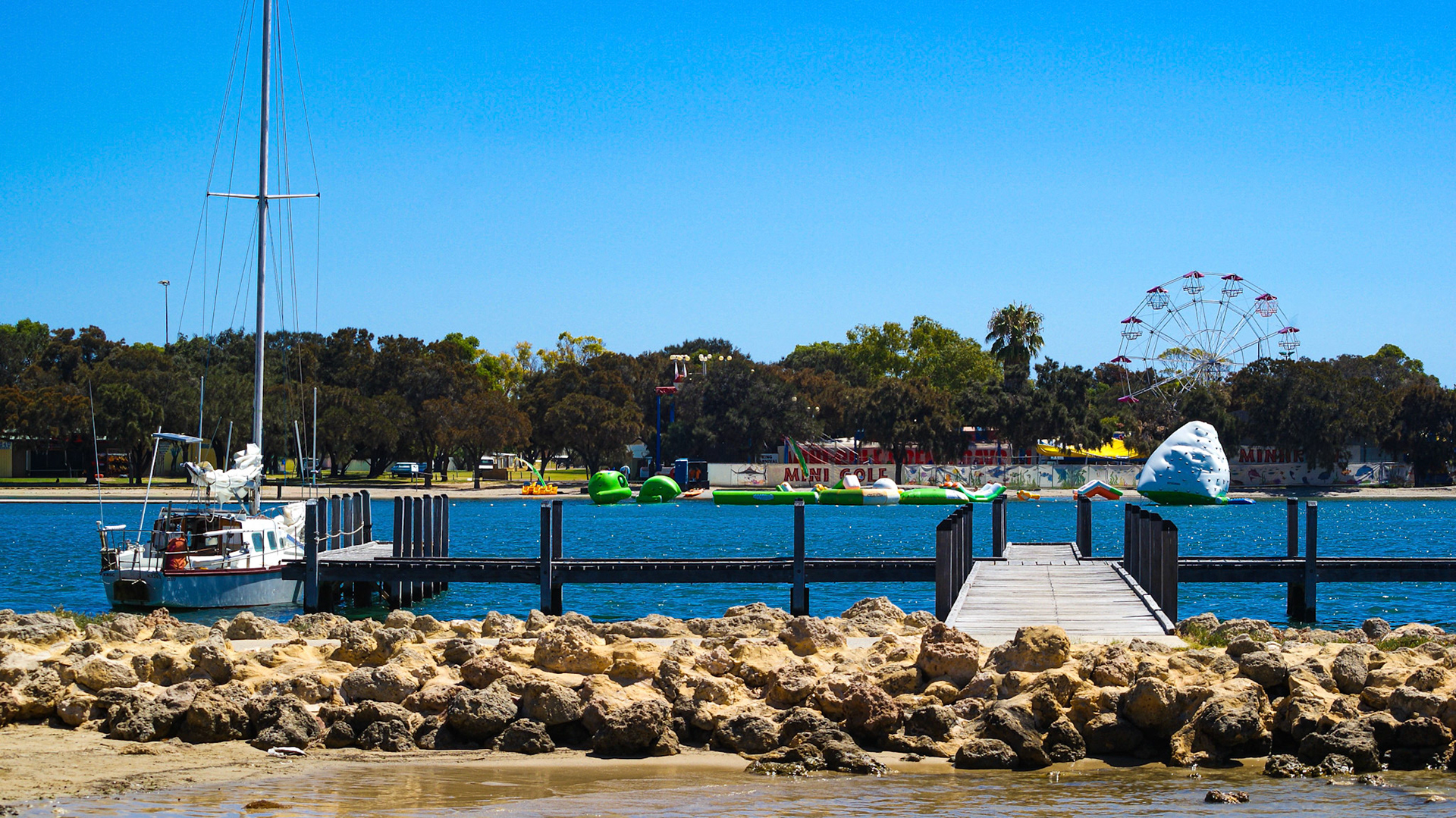 A sunny waterfront scene with a wooden jetty stretching over calm blue water. A sailboat is moored to the left, while bright green inflatables and a white climbing “iceberg” float form part of a water park across the channel. Behind the trees, an old-style Ferris wheel and a mini-golf area add a playful backdrop. Rocky shoreline and a clear blue sky complete the lively coastal view.