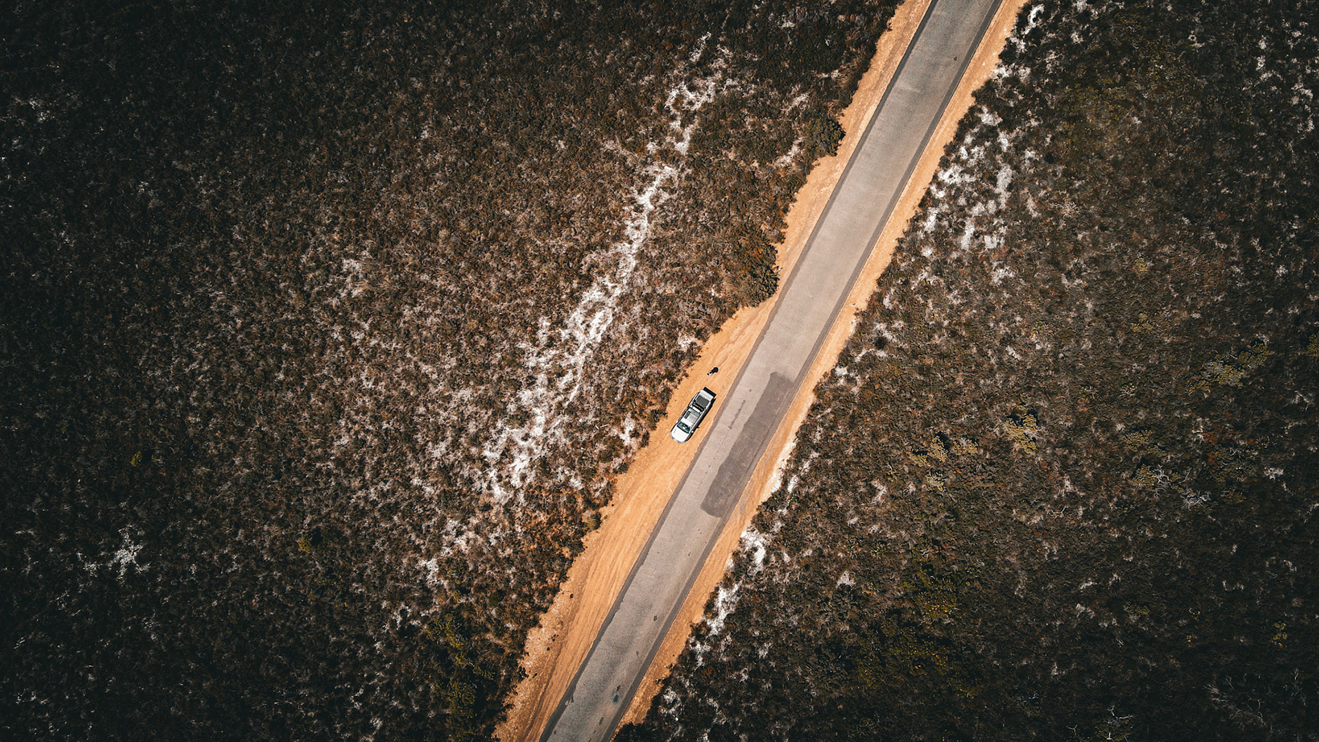 An aerial view shows a solitary car parked on an asphalt road in a vast, arid landscape, evoking themes of solitude and exploration.