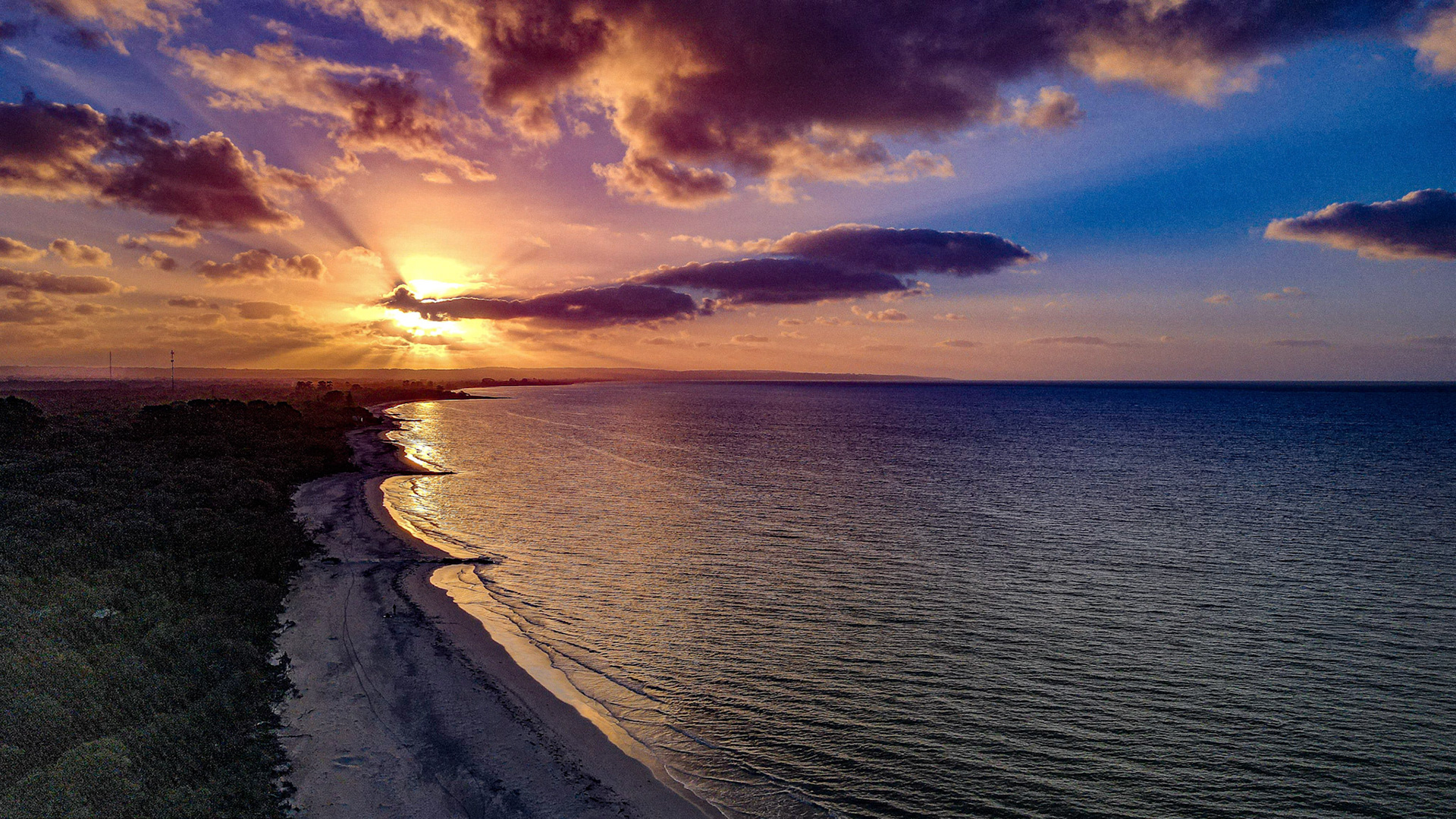 Stunning aerial view of a serene sunset over a tranquil coastline. The vibrant colors of the sky blend beautifully as the sun sets, casting a warm glow over the ocean and beach. Gentle waves lap against the shore, creating a peaceful and idyllic scene perfect for capturing the beauty of nature.