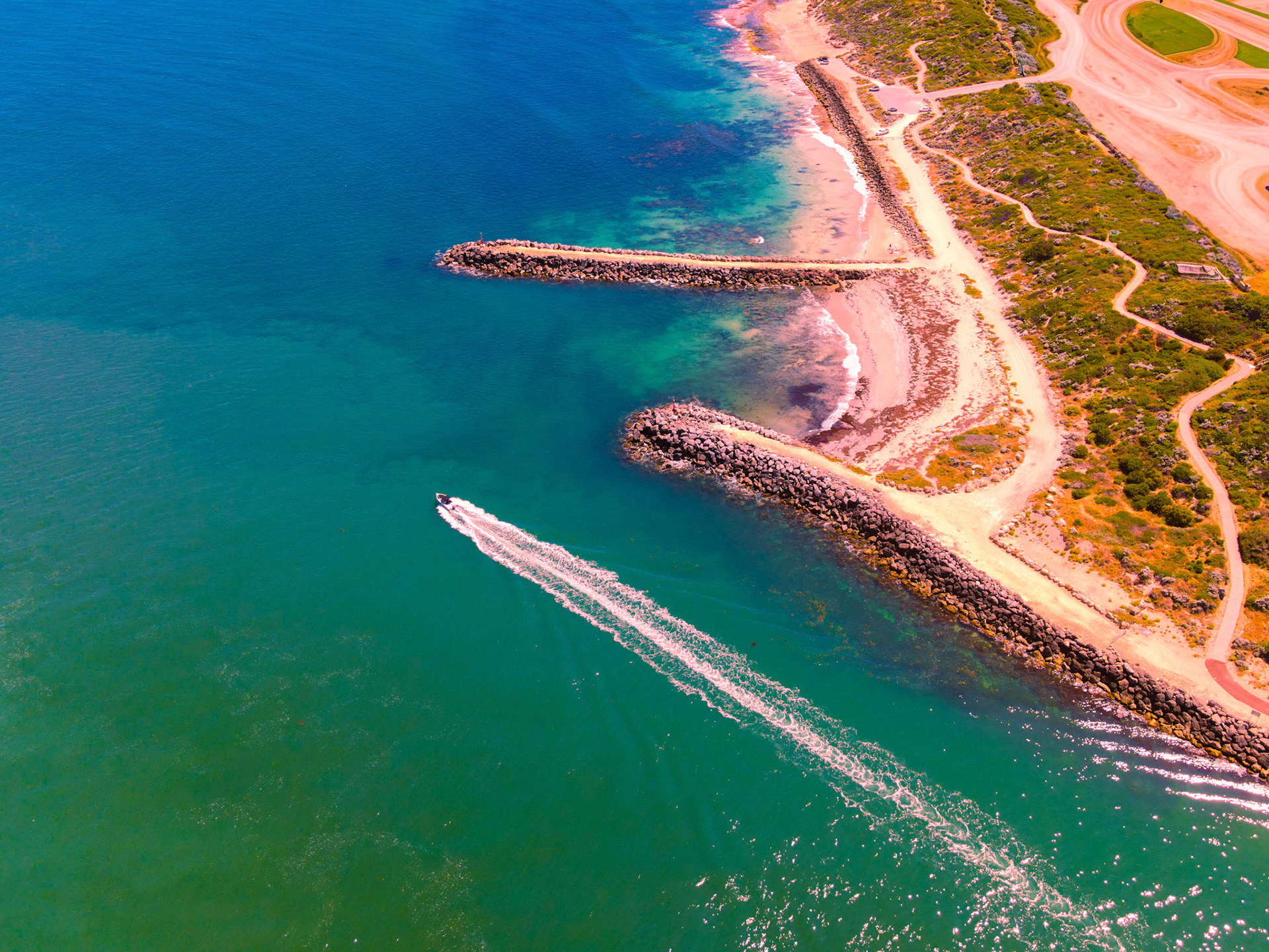 Vibrant aerial shot of a sandy headland with two curved rock breakwaters extending into clear turquoise water. A small motorboat cuts across the sea, leaving a long white wake behind it. Sunlit shallow areas reveal patches of seagrass and changing water tones from teal to deep blue. Winding coastal paths and low scrub cover the shoreline, and the sandy beach curls around the protected inlet.