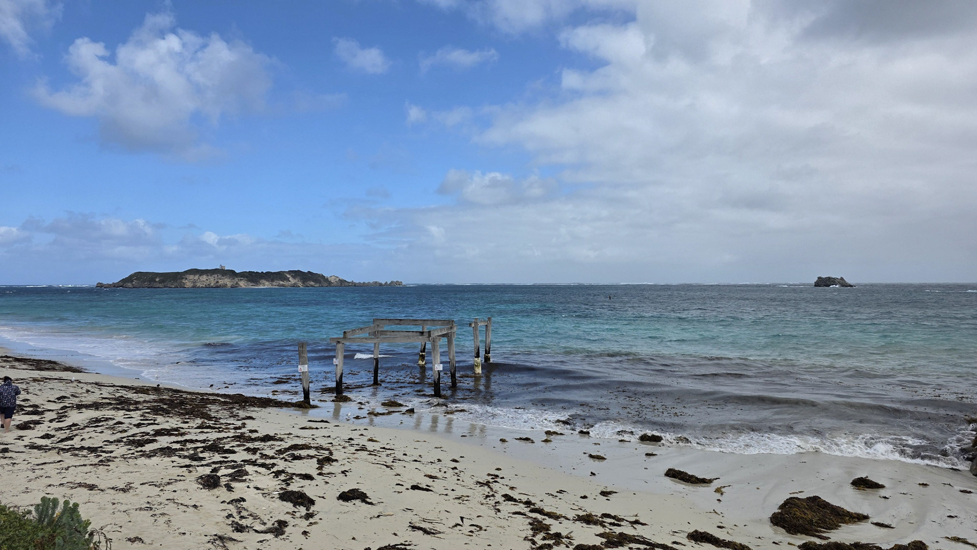 A tranquil coastal scene depicting the remains of a wooden pier stretching into the turquoise sea. The sandy beach, scattered with seaweed, extends under a partly cloudy sky, creating a peaceful and natural ambiance. In the distance, an island and rocky outcropping enhance the picturesque maritime setting.