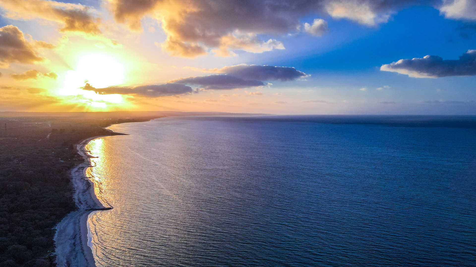 A breathtaking aerial view capturing the vibrant hues of a sunset over a tranquil ocean. The sun casts a golden glow across the water, reflecting off gentle waves, while fluffy clouds add depth to the warm, colorful sky. The coastline curves gracefully, offering a peaceful and inspiring scene that evokes a sense of calm and wonder.