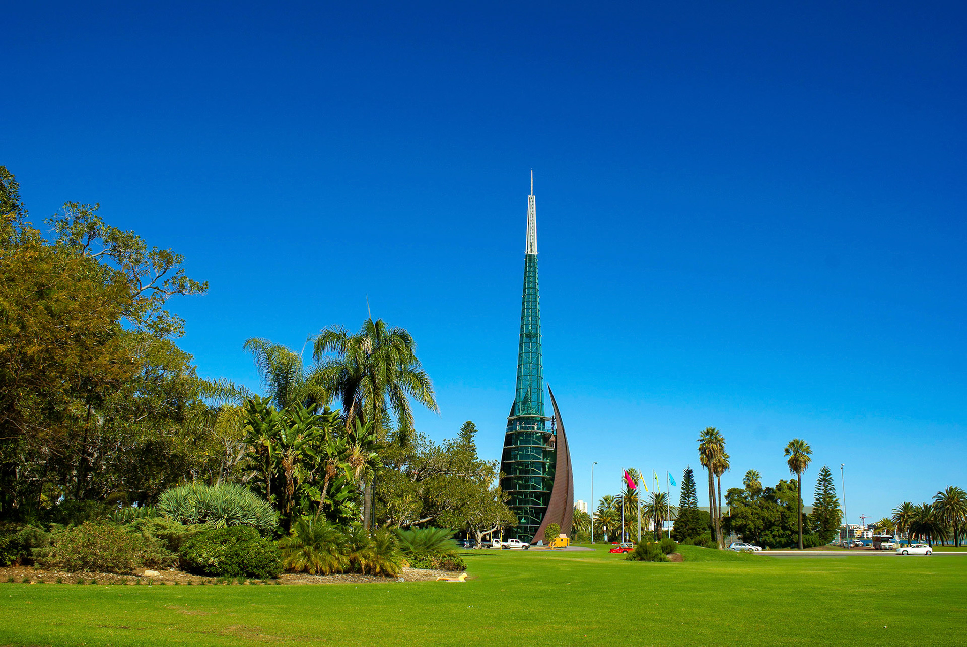 A striking modern spire stands tall against a clear blue sky, surrounded by lush green grass and trees in a vibrant urban park.