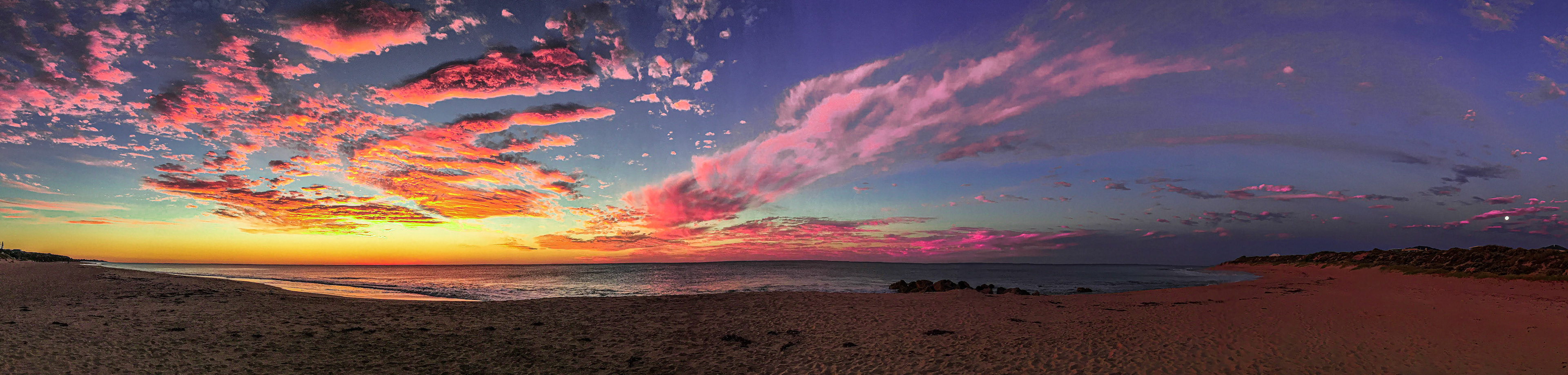 A panoramic view showcases a stunning sunset over the ocean, with vibrant colors reflecting on the calm sea and sandy beach.