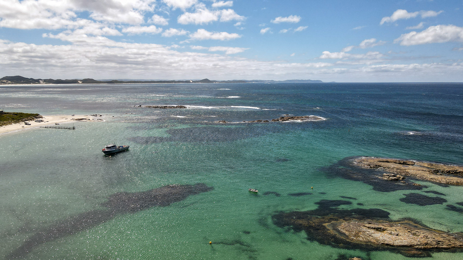 An aerial view showcases a tranquil coastal landscape with clear waters, a boat anchored in the bay, and people on the sandy beach.