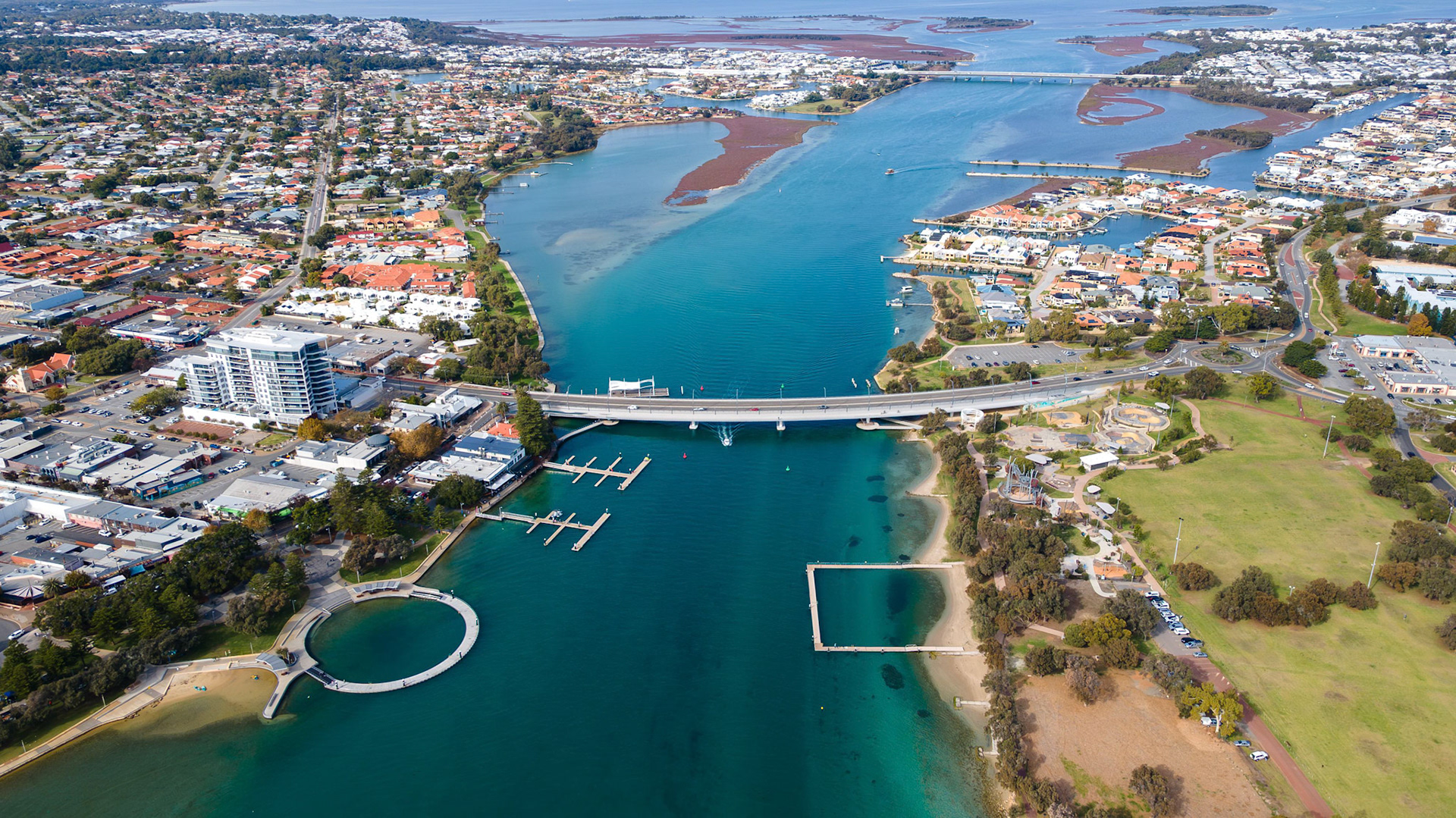 This vibrant aerial shot captures a beautiful coastal city with a serene waterway, showcasing a blend of natural beauty and urban development. The image features a winding river lined by attractive residential areas and commercial properties, with a prominent bridge connecting different sections of the city. The shot exhibits vibrant colors and clear water, creating a visually appealing scene that highlights the harmony between nature and human habitation. Ideal for use in travel brochures, city promotional materials, or websites focusing on urban development and nature tourism.